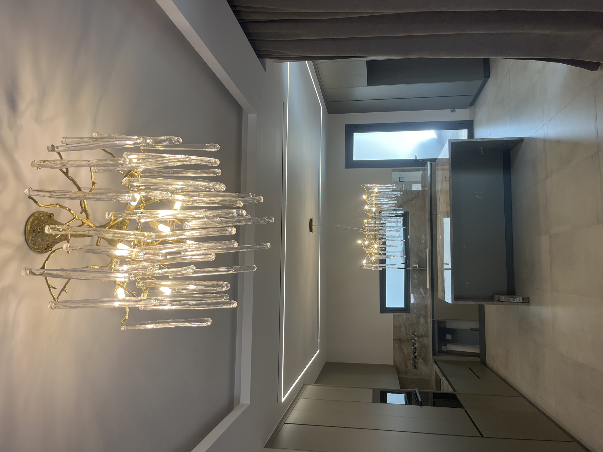 This is a full-shot, eye-level, interior photograph of a modern kitchen in Moni Community, Cyprus. The kitchen features a large island with a grey countertop, custom cabinetry in a muted olive green, and a sleek marble backsplash. Two prominent chandeliers, one hanging above the island and another suspended from the ceiling, illuminate the space with a warm glow. The room also includes recessed ceiling lights and a large window offering a view of the outdoors. A tall, dark-framed door is visible in the background, leading to an area with natural light. The floor is tiled in a neutral tone. The photograph is well-lit and captures the contemporary design elements of the kitchen.