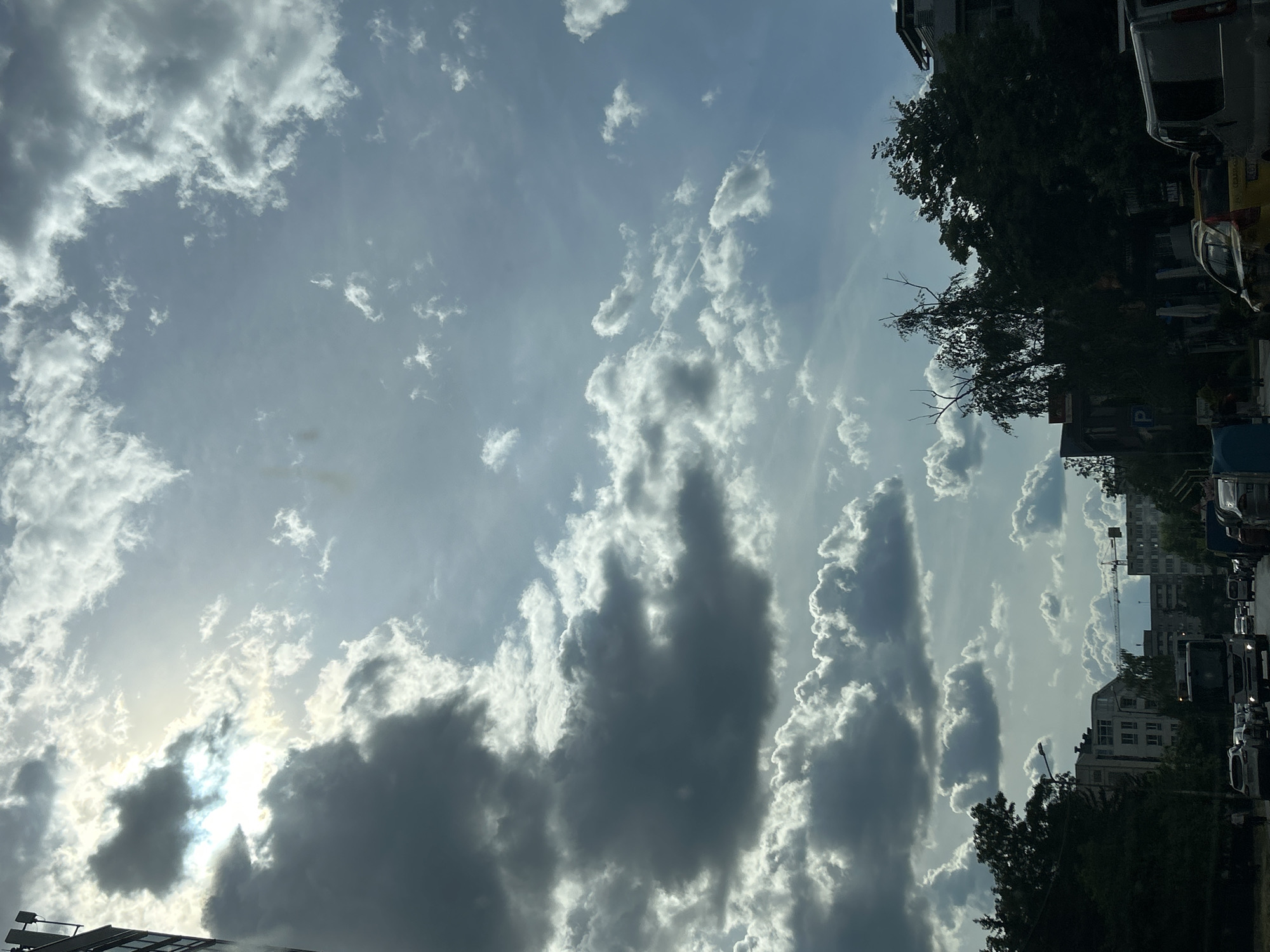 The image shows a view of the sky with numerous clouds, some dark and others lighter, with the sun shining through them. The scene includes the tops of buildings and trees along a street, suggesting an urban setting. Cars are parked along the side of the street. The location is Sofia, Bulgaria, as evidenced by local signage in Bulgarian. There are no people visible in the image. A crane can be seen behind the buildings.