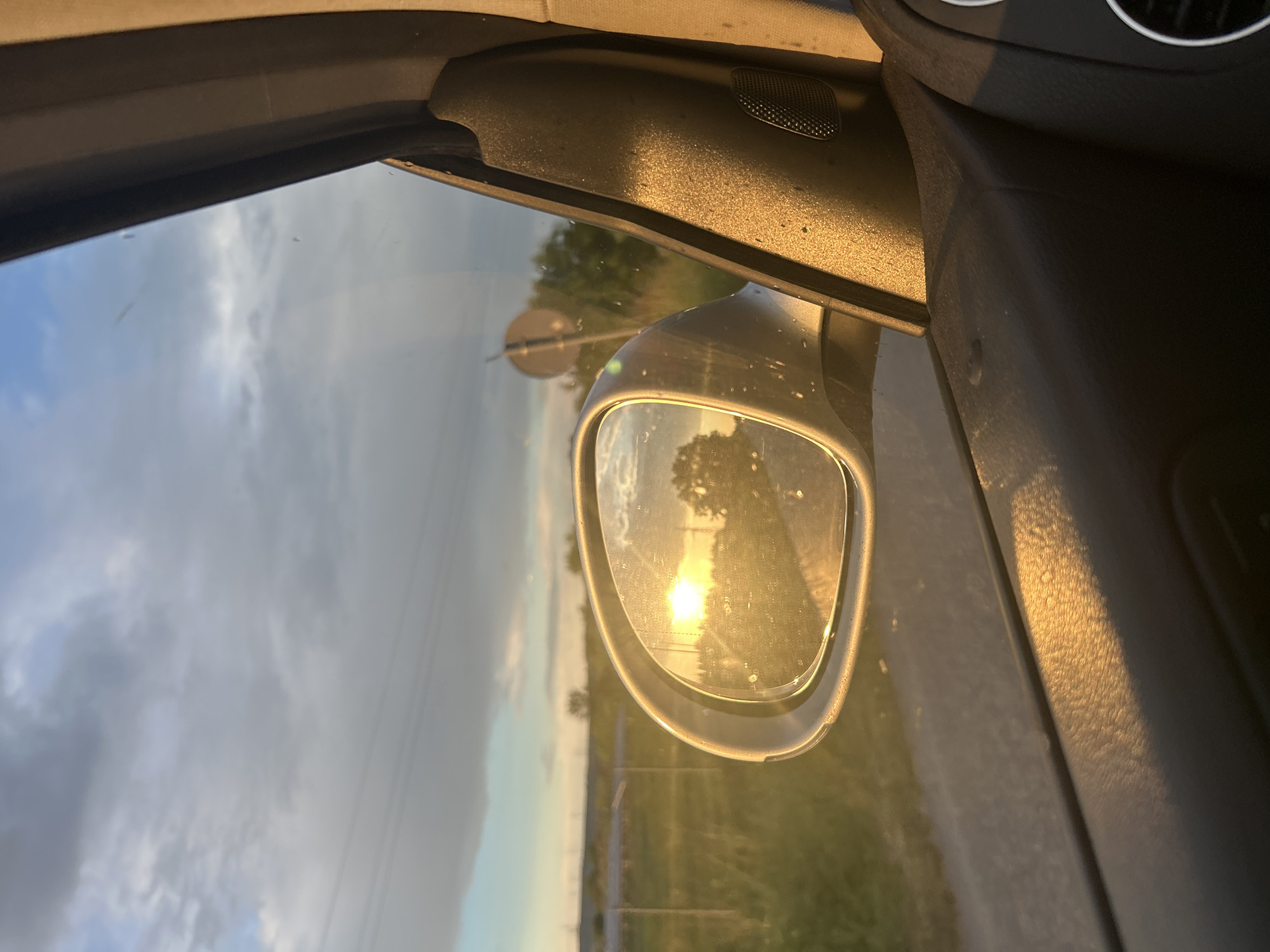 The scene depicts a view from the interior of a vehicle, looking through the front passenger side window towards the exterior in Milkovitsa, Bulgaria, during a late afternoon or early evening period. The sun, appearing as a bright, low-angle golden light source, is directly reflected within the silver-gray exterior side mirror, causing a lens flare effect and illuminating the mirror's surface with intense glare. The mirror exhibits minor surface imperfections or dust.
The immediate foreground on the right consists of the dark, textured plastic interior door panel of the vehicle, which is partially illuminated by the warm, golden sunlight. A small, dark speaker grille is visible on the door panel. A section of the car's window frame and a portion of the dashboard are also visible on the far right. The window itself shows minor streaks or dust.
Outside the vehicle, a paved, dark gray road surface is partially visible, paralleling the vehicle on the left. Adjacent to the road is an uncultivated area characterized by green and yellowing tall grass and sparse vegetation. In the mid-ground, beyond the grassy area, several vertical elements resembling fence posts or utility poles are present. Further into the mid-ground, a circular road sign, dark in color and with an indistinct symbol, stands against a backdrop of distant trees.
The sky above the horizon is largely covered by a formation of stratocumulus or nimbostratus clouds, appearing in shades of gray and white. Patches of lighter blue sky are visible between the cloud formations, particularly in the upper left quadrant. Thin, dark horizontal lines, consistent with power lines, are faintly visible against the sky in the upper left. No individuals are visible in the observable environment.