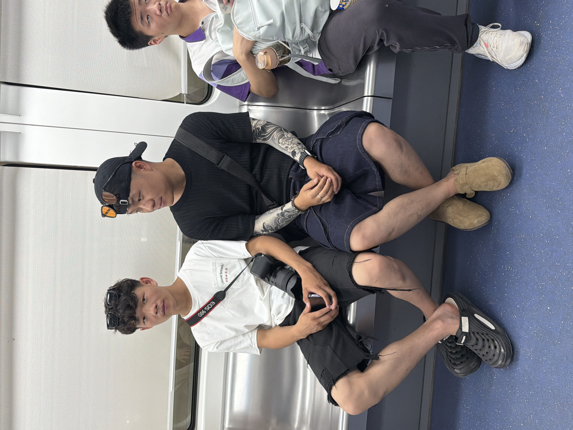 Three young East Asian males are seated on a silver bench inside a subway car, likely in Yuzhong District, China. The person on the left, wearing a white t-shirt and black distressed shorts, has sunglasses on his head and a camera strap around his neck with a DSLR camera attached. The middle person wears a black t-shirt, a black baseball cap with sunglasses perched on top, and dark denim shorts. His arms are adorned with large, detailed tattoos of floral designs. The person on the right, wearing a purple and white polo shirt and dark grey pants, is partially visible and holds a light blue backpack. The subway car interior features a blue floor and white roller blinds in the background.