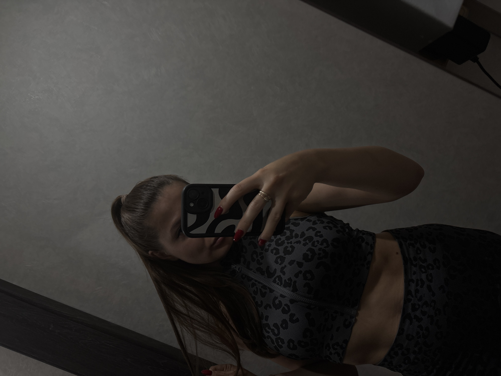 A young woman with a high ponytail and dark hair is taking a selfie in a mirror. She is wearing a leopard-print sports bra and matching bottoms. Her nails are painted red. She is holding her phone, which has a black and white patterned case, in her right hand. Her left hand is pulling on her hair. The background is a gray wall. The scene is dimly lit.