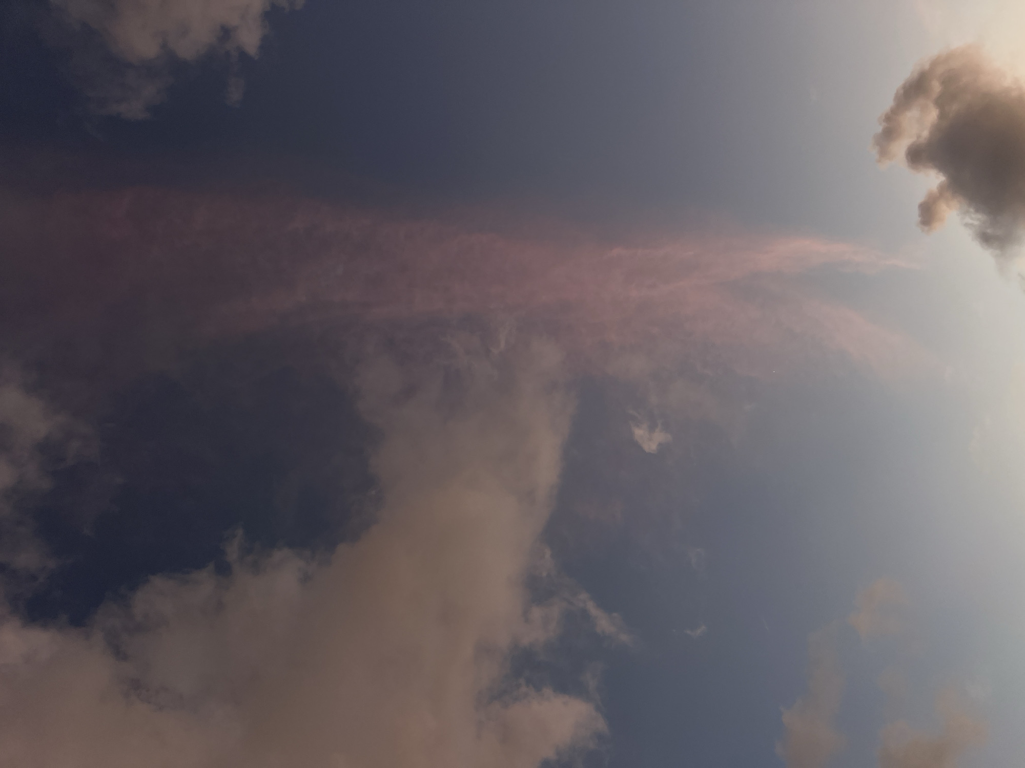The image shows a cloudy sky in Choeng Thale, Thailand. The sky is a gradient of deep blue at the top to a lighter blue towards the horizon. There are several clouds visible. A prominent wispy cloud formation with a pinkish hue extends vertically in the center of the image. To the left, there are larger, more diffused clouds with a warm, possibly sunset-induced, coloration. In the lower right corner, a darker, more defined cloud mass is visible. The overall impression is of a sky during dusk or dawn, with dramatic cloud formations. No people, structures, or specific landmarks are identifiable.