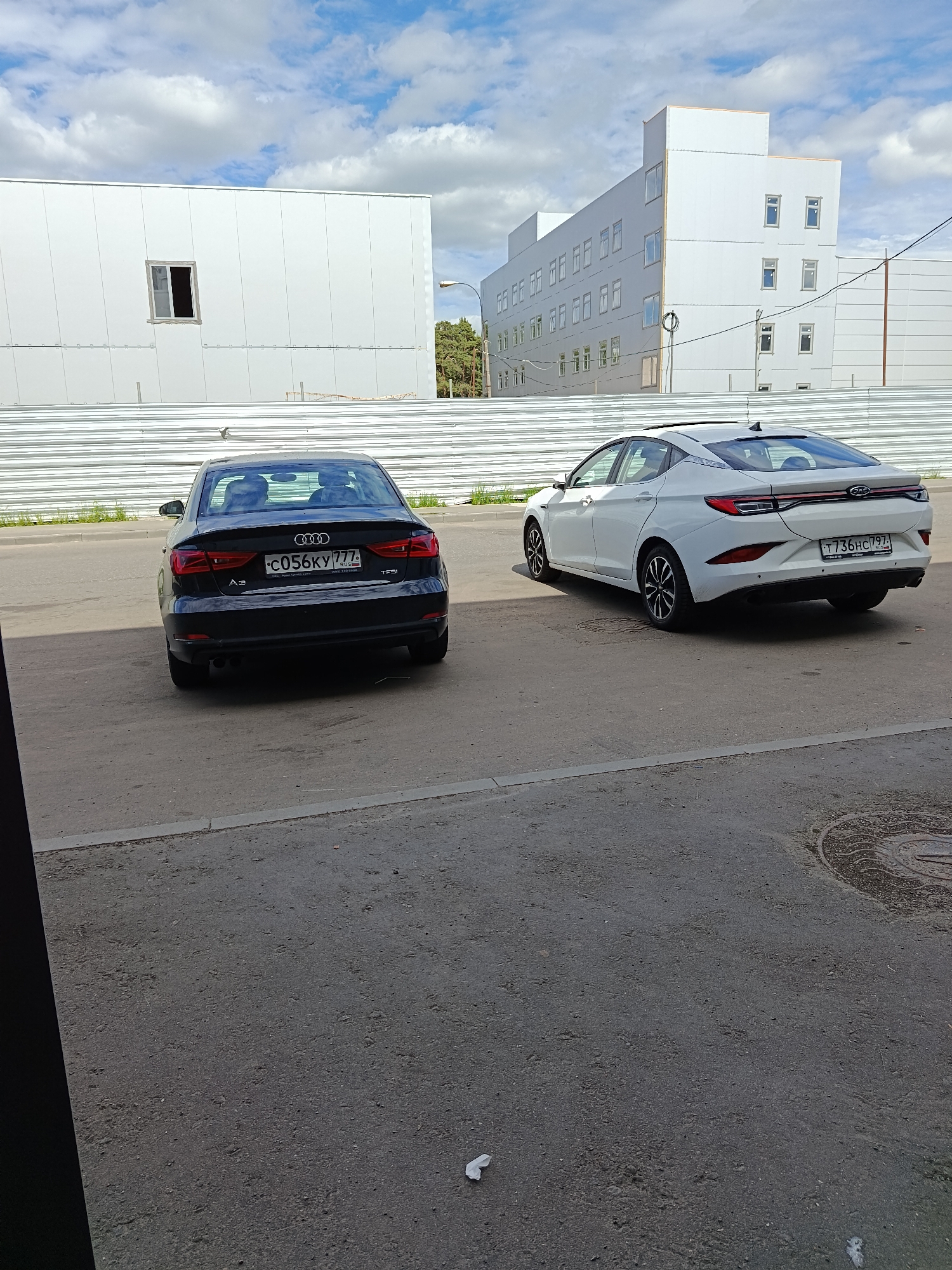 The image shows two cars parked on a paved road in Moscow, Russia. The car on the left is a black Audi A3 with the license plate C056KY777. The car on the right is a white JAC J7 with the license plate T736HC797. In the background is a construction fence, and behind it, two white buildings are visible. The sky is partly cloudy. The location appears to be an industrial or commercial area.