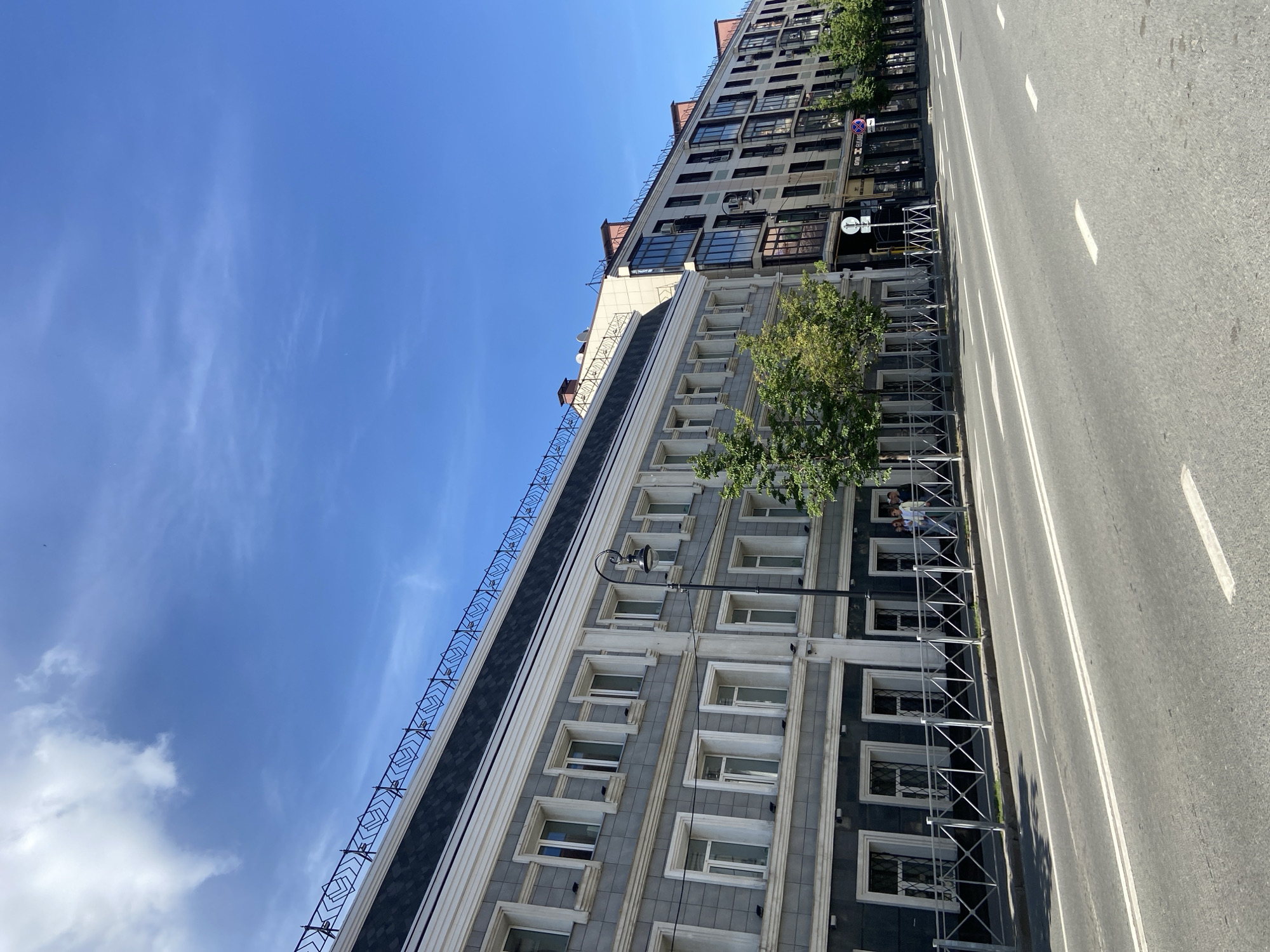 The image depicts an urban street scene on a bright, sunny day in Kazan, Russia, specifically along Pushkin Street. The view is from across a multi-lane asphalt road looking towards a row of buildings on the opposite side.
The sky is a clear, vibrant blue with scattered wisps of white clouds, primarily on the upper left. The road, devoid of vehicles, has two prominent double white lines marking lanes and several dashed white lines. A dark gray sidewalk runs parallel to the road, separated by a light gray metal pedestrian barrier with intersecting diagonal supports.
On the left, a multi-story building with a gray facade dominates, featuring numerous white-framed windows with dark sills and white decorative trim. The building has a prominent mansard-style roof with dark shingles and an ornate black metal railing forming geometric patterns along its edge. A black street lamp with a single light fixture stands in front of this building. Several small, green deciduous trees are planted along the sidewalk. A group of three to four casually dressed individuals is visible on the sidewalk in front of the gray building, seemingly walking or standing near an entrance. One person is wearing a light-colored top and dark pants, another is in a dark shirt and pants.
To the right of the gray building, a more modern, taller building extends down the street. It features a lighter, beige or off-white facade with many dark-framed windows and balconies. Visible on its roof are several antennas and chimneys. Commercial signs are partially visible on the ground level of this building, including one clearly displaying "БАНК" (Bank) and another appearing to say "ЕЛІСАВ" or "ELISABETH" next to it. No distinct interactions are visible between the people or with the environment beyond their presence on the sidewalk.