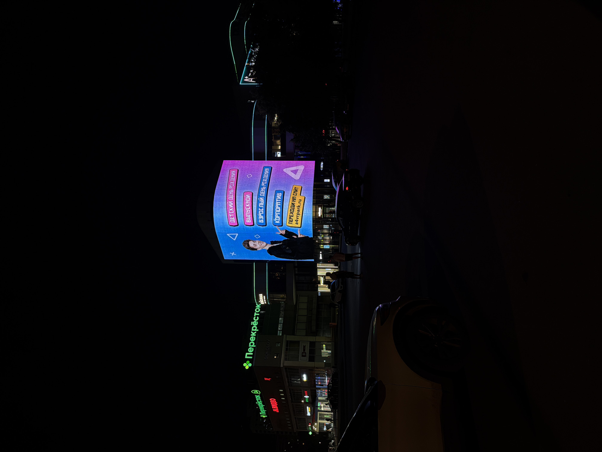 This image captures a nighttime urban scene in Moscow, Russia, specifically located within the Moscow International Business Center area, likely near AFI Mall City. The dark sky is illuminated by numerous artificial lights from buildings and a prominent digital billboard.
On the left, a multi-story commercial building features several illuminated signs: multiple green "Перекрёсток" (Perekryostok) supermarket logos, a red "M.BИДЕО" (M.Video) electronics store sign, a "SOKOLOV" jewelry sign, and a "MEZANA" sign. An "Аптека" (Pharmacy) sign is also visible at street level.
In the center-right, a large, vertically oriented digital billboard displays a vibrant blue and pink advertisement. It features a smiling man in a dark jacket, gesturing with both hands. The billboard's text, in Russian, promotes various events: "ДЕТСКИЙ ДЕНЬ РОЖДЕНИЯ" (Children's Birthday), "ВЫПУСКНОЙ" (Graduation), "ВЗРОСЛЫЙ ДЕНЬ РОЖДЕНИЯ" (Adult's Birthday), and "КОРПОРАТИВ" (Corporate Event). A yellow banner at the bottom prompts "ПЕРЕХОДИ НА САЙТ a4vrpark.ru" (Go to the website a4vrpark.ru), indicating an advertisement for a VR park.
To the right of the digital billboard, another multi-story building is partially visible, distinguished by green neon lighting tracing its roofline and a prominent "A+" logo.
In the foreground, the front-left section of a light-colored SUV is visible. On the street, two individuals stand near the base of the digital billboard, and a dark sedan is present, along with other vehicles further in the distance. The scene conveys a dynamic, brightly lit commercial district after dark.