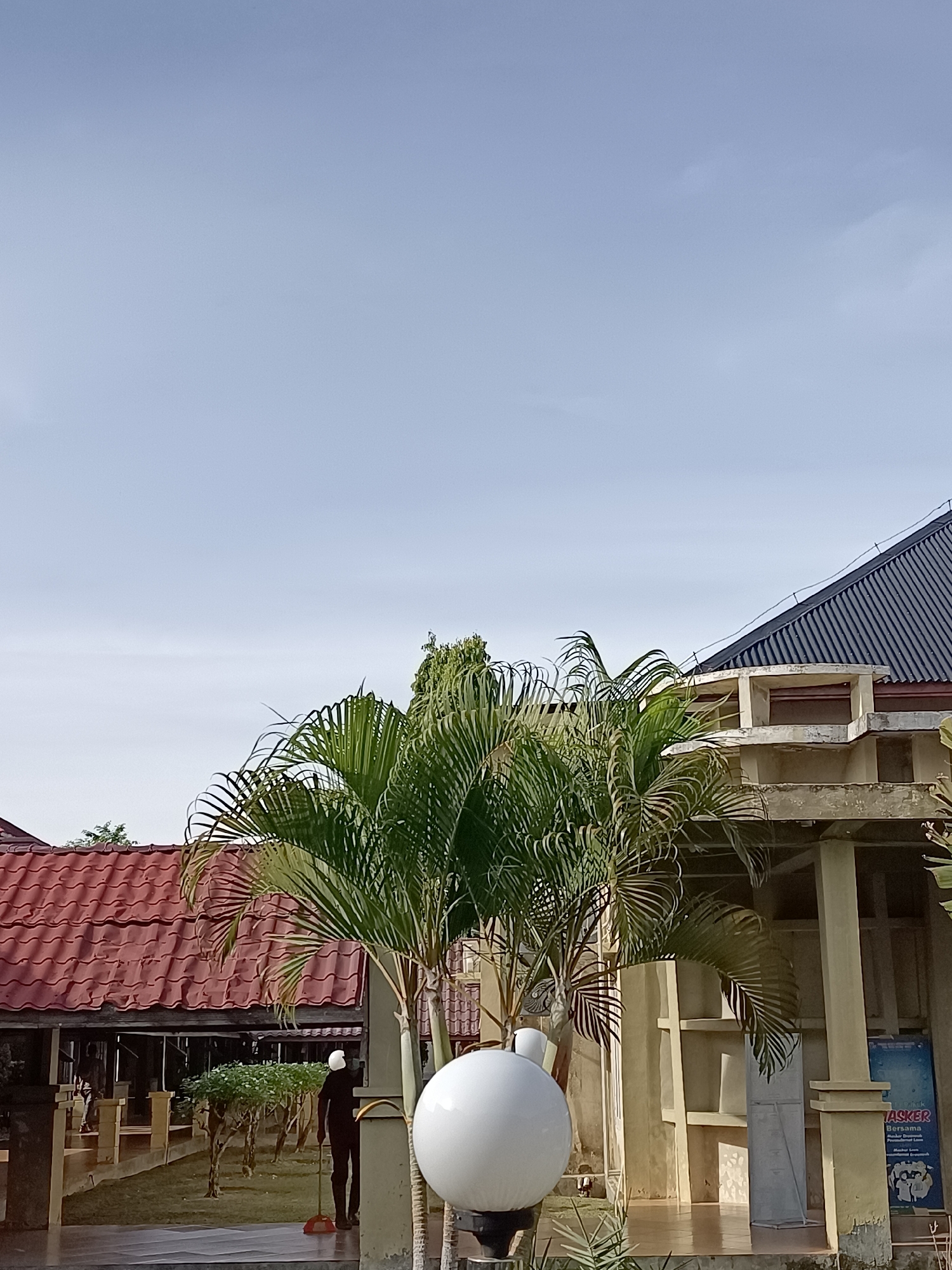 In Mesjid Reuntoh, Indonesia, a bright, clear sky dominates the upper portion of the image. Below, palm trees with lush green fronds are positioned in the foreground and middle ground. To the left, a building with a red tiled roof is visible, featuring a covered walkway with supporting pillars. To the right, a building with a dark, sloped roof and concrete structural elements is present. A person dressed in dark clothing is seen standing near a broom and dustpan, appearing to sweep the ground. In the immediate foreground on the right, a large, round, white light fixture is positioned. A blue sign with text, including the word "MASKER," is attached to the right-hand building. The environment appears to be an outdoor area, possibly a courtyard or garden, adjacent to the buildings.