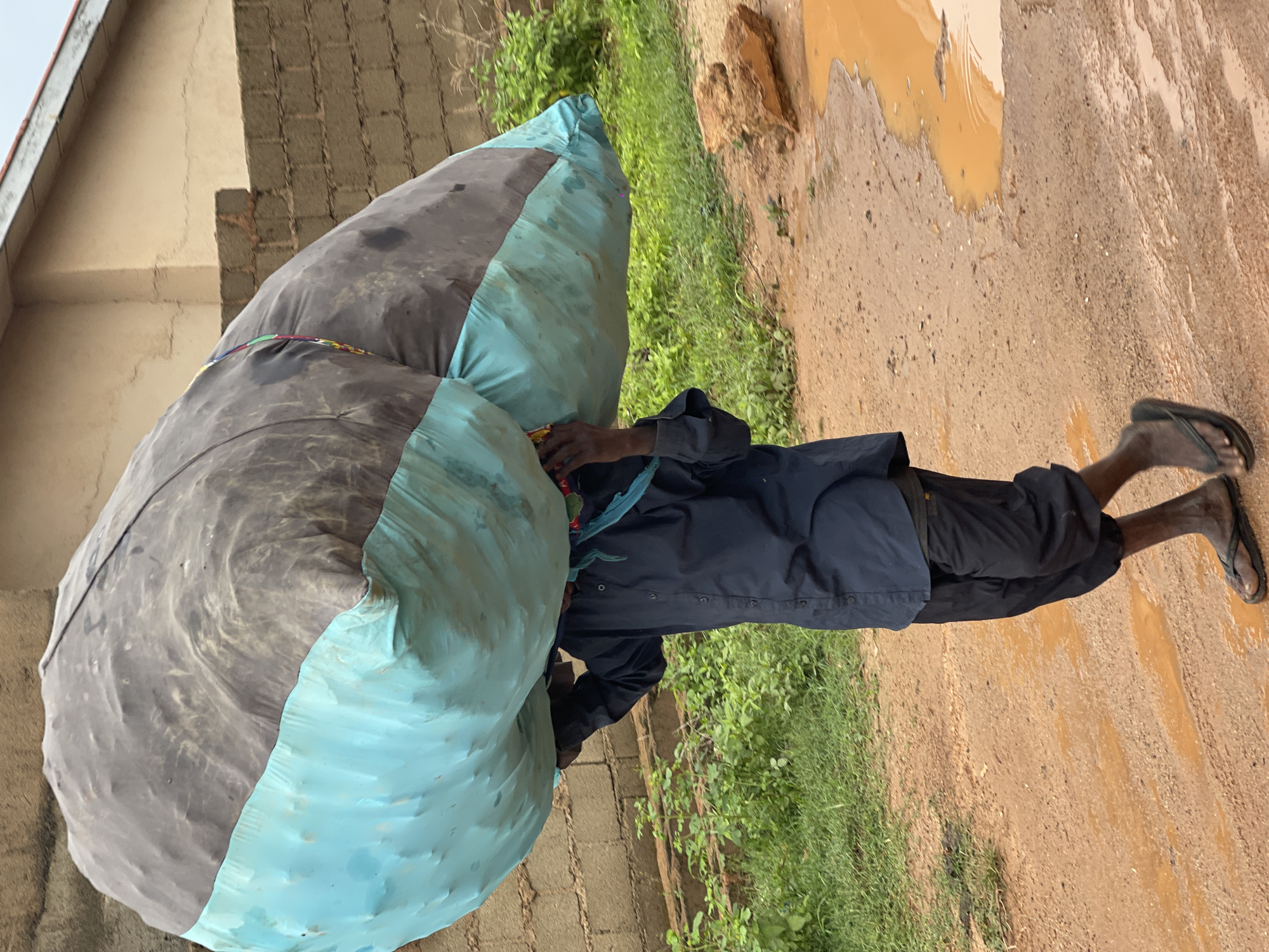A person wearing a dark blue button-down shirt and dark pants walks on a dirt road in Jos, Nigeria. They are carrying a very large, lumpy bundle on their shoulders, which appears to be covered in a blue tarp. The bundle is secured with straps or ropes, and there is a colorful piece of fabric visible at the top of the bundle. The road is muddy, with several puddles of water. To the left of the road, there is green grass and some sparse vegetation. In the background, there is a brick wall and part of a building. The sky is overcast.