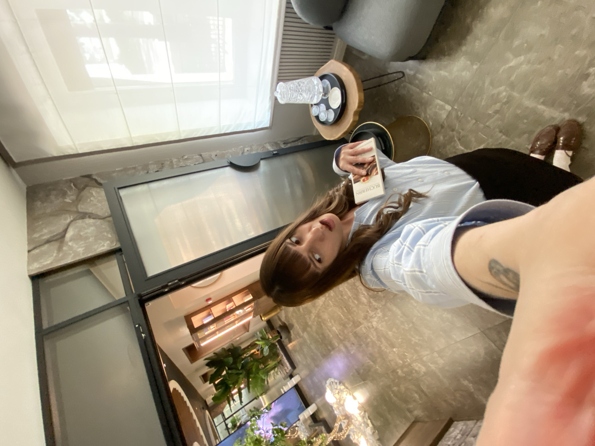 The image shows a woman holding a book with the word "BUCHERON" on the cover. She is wearing a light blue striped shirt and a dark skirt. Her shoes are visible, consisting of white socks and brown dress shoes. She is standing indoors on a gray patterned floor. A small wooden table with glassware is near her. Behind her, a large window reflects the interior of what seems to be a shop or a display area with plants and lighting fixtures. The image's composition is such that the foreground is slightly blurred, suggesting a close-up shot. The location is impossible to determine based on the image alone.