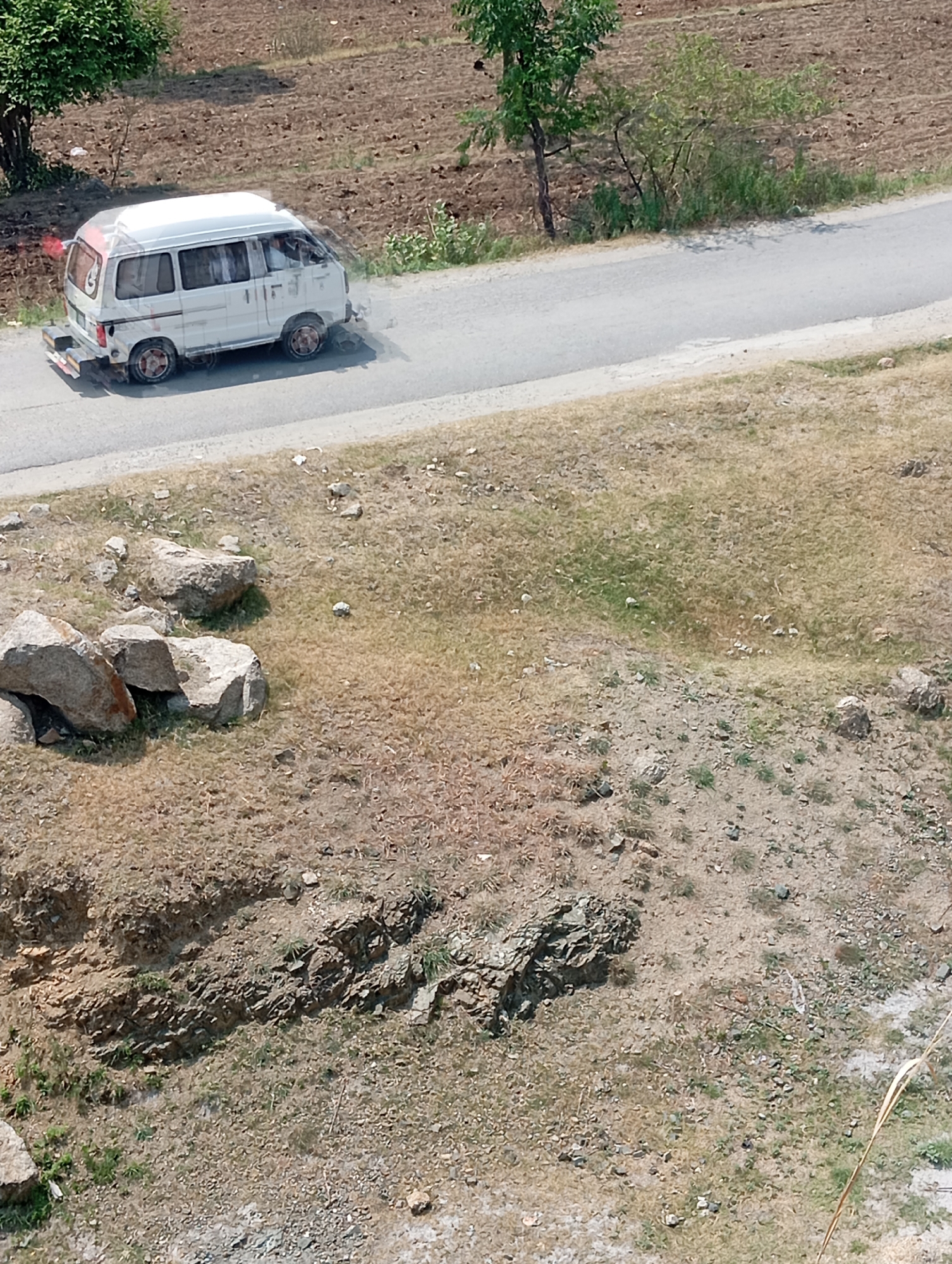 In Hakale, Pakistan, a white van is seen traveling on a paved road. The van appears to be an older model, possibly a microvan, with tinted windows. A hazy cloud of exhaust is visible behind the vehicle, indicating its movement. The road itself is in good condition, with a clear lane marking visible. The surrounding environment consists of dry, grassy terrain with scattered rocks and some sparse vegetation. In the background, a cultivated field can be seen, suggesting agricultural activity in the area. The scene appears to be a typical roadside view in a rural or semi-rural setting within Pakistan.
