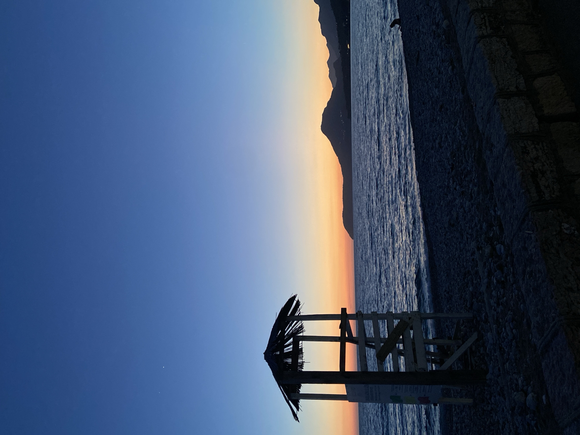 The image depicts a dusk coastal scene on a pebbly beach in Bar, Montenegro. The sky transitions from a deep indigo blue overhead to a band of orange and yellow hues at the horizon line. Two distinct, small white points of light are visible in the upper-left quadrant of the sky. The sea surface is dark blue-purple, displaying subtle wave patterns. In the left midground, a wooden lifeguard tower with a distinctive thatched roof stands in silhouette. A light-colored rectangular sign is affixed to the left support of the tower. Along the horizon in the background, a series of dark, undulating mountains are silhouetted, with small clusters of artificial lights discernible at their base on the far right, indicating coastal development. In the far right midground, a solitary human figure is seated on the pebbly beach near the water's edge, also rendered in silhouette. A low, stacked stone wall, composed of rough-hewn blocks, occupies the immediate foreground.