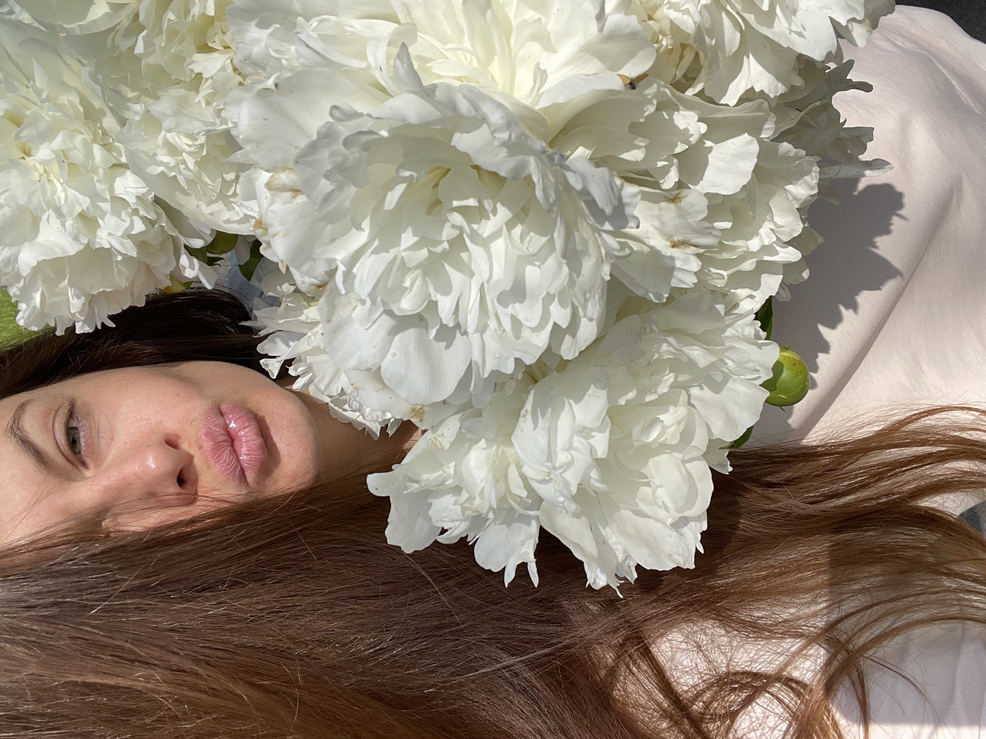 This media file captures a close-up, overhead view of a person lying down amidst a large bouquet of white peonies. The person's face is partially visible, turned towards the camera, with their eyes open and lips slightly parted. Their long, brown hair spreads across the scene, partially obscuring the peonies and resting on a light-colored fabric. The peonies are abundant, with many large, ruffled white petals, some showing slight discoloration or water droplets, suggesting they are fresh. The lighting indicates it is daytime, with strong sunlight casting shadows from the flowers onto the fabric and the person's hair, suggesting an outdoor setting or a brightly lit indoor space. The overall mood is serene and artistic, with a focus on natural beauty and texture. There is no visible text in the image. The location context provided is Zarechny, Russia.