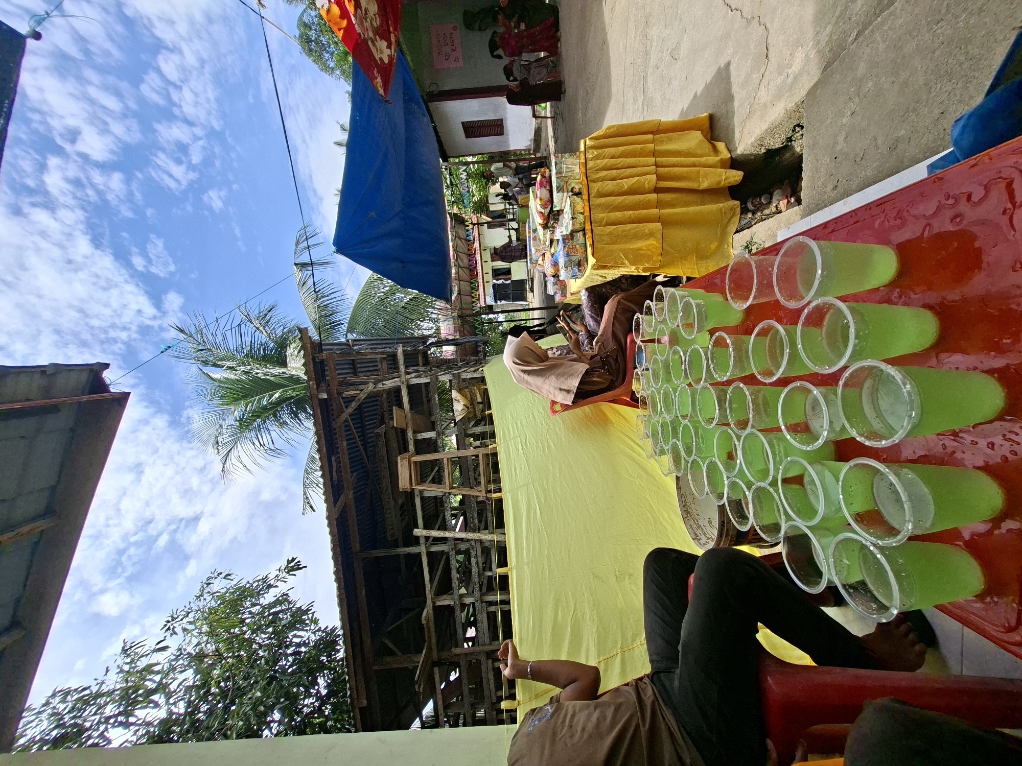 In Kruengmate, Indonesia, an outdoor gathering is taking place under a bright blue sky with scattered clouds. The scene is set in a partially covered area, possibly a community event or informal celebration. In the foreground, a red table displays numerous plastic cups filled with a pale green liquid, presumably a beverage. A person in a brown t-shirt and dark pants is seated on a red stool to the left, facing away from the camera, with their left arm resting on their leg. To the right of the drinks table, a woman wearing a hijab and a brown dress is seated, looking at her phone. Behind them, a yellow tarp is stretched across a wooden structure, with more people and activity visible in the background. A blue tarp is also visible, providing additional shade. Further in the background, buildings and trees suggest a rural or semi-rural setting. A table covered with a yellow tablecloth is laden with boxes, possibly gifts or food items, and several people are gathered around it. Children in colorful clothing are also present in the background, interacting with adults. The overall impression is one of a casual community event, with refreshments and social interaction.