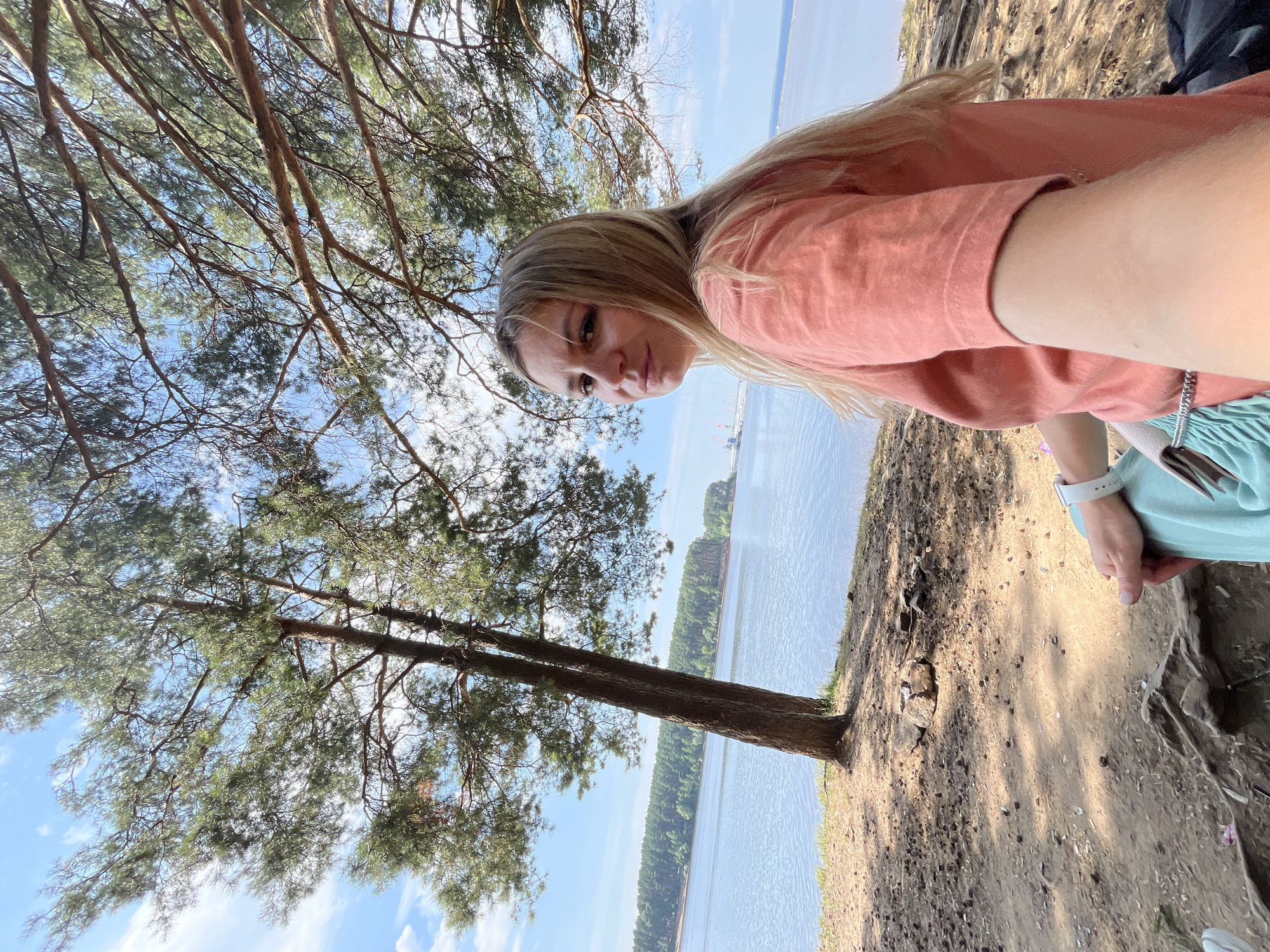 A blonde woman sits on the ground under the branches of a pine tree, looking at the camera. She is wearing a coral-colored t-shirt and a teal skirt. The tree is located on the shore of the Kama River in Chaikovsky, Russia. The river stretches out behind her, with a tree-lined shore on the opposite bank. The sky is blue with scattered white clouds. The ground is covered with dirt, pine needles, and small stones. The scene appears to be a peaceful, natural setting.