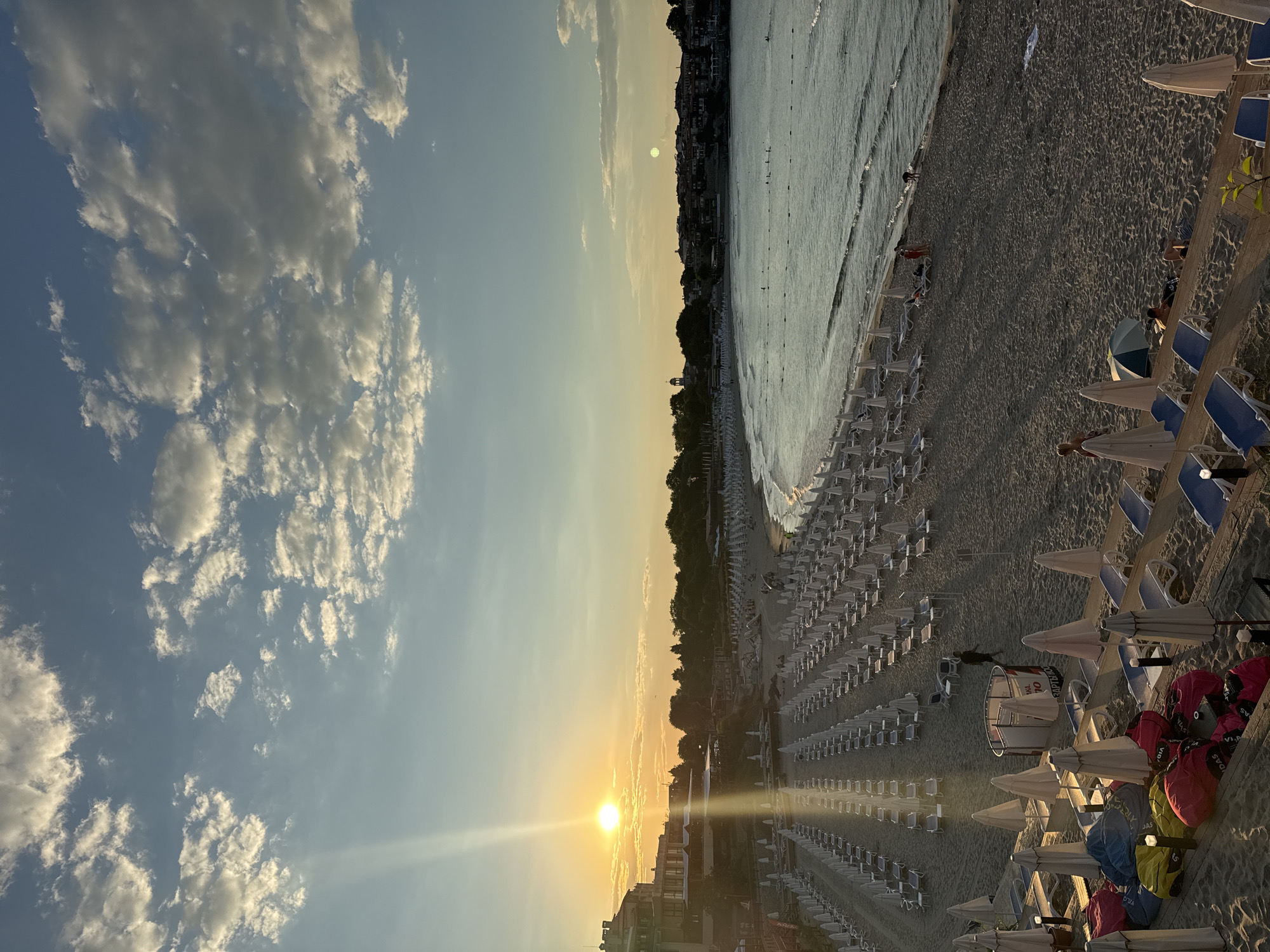 The image captures a panoramic view of a beach in Sozopol, Bulgaria, during sunset. The sun, positioned low on the horizon, casts a warm, golden light across the scene, with visible sunbeams fanning out into the sky. The sky is a blend of pale blue and soft yellow, dotted with scattered white and gray clouds.
The foreground of the beach is filled with rows upon rows of neatly arranged beach chairs and umbrellas, most of which are closed and white. Some colorful beanbag chairs are also visible in the lower left portion of the frame. A few people are scattered across the sand, some walking and others lounging on chairs or by the water's edge. One individual in a red dress is standing near the water, and another person is bent over by the shoreline.
The middle ground shows the gentle waves of the sea meeting the sandy shore. A few people are swimming or wading in the water. The coastline curves gently, and beyond the beach, a town with buildings featuring red-tiled roofs and some greenery is visible. The architecture suggests a historical European coastal town. Trees line the edge of the town.
In the distance, the horizon is marked by the silhouette of the town and the sea merging with the sky. The overall atmosphere is serene and peaceful, typical of a late afternoon on a vacation spot. The lighting conditions indicate that it is either sunrise or sunset.