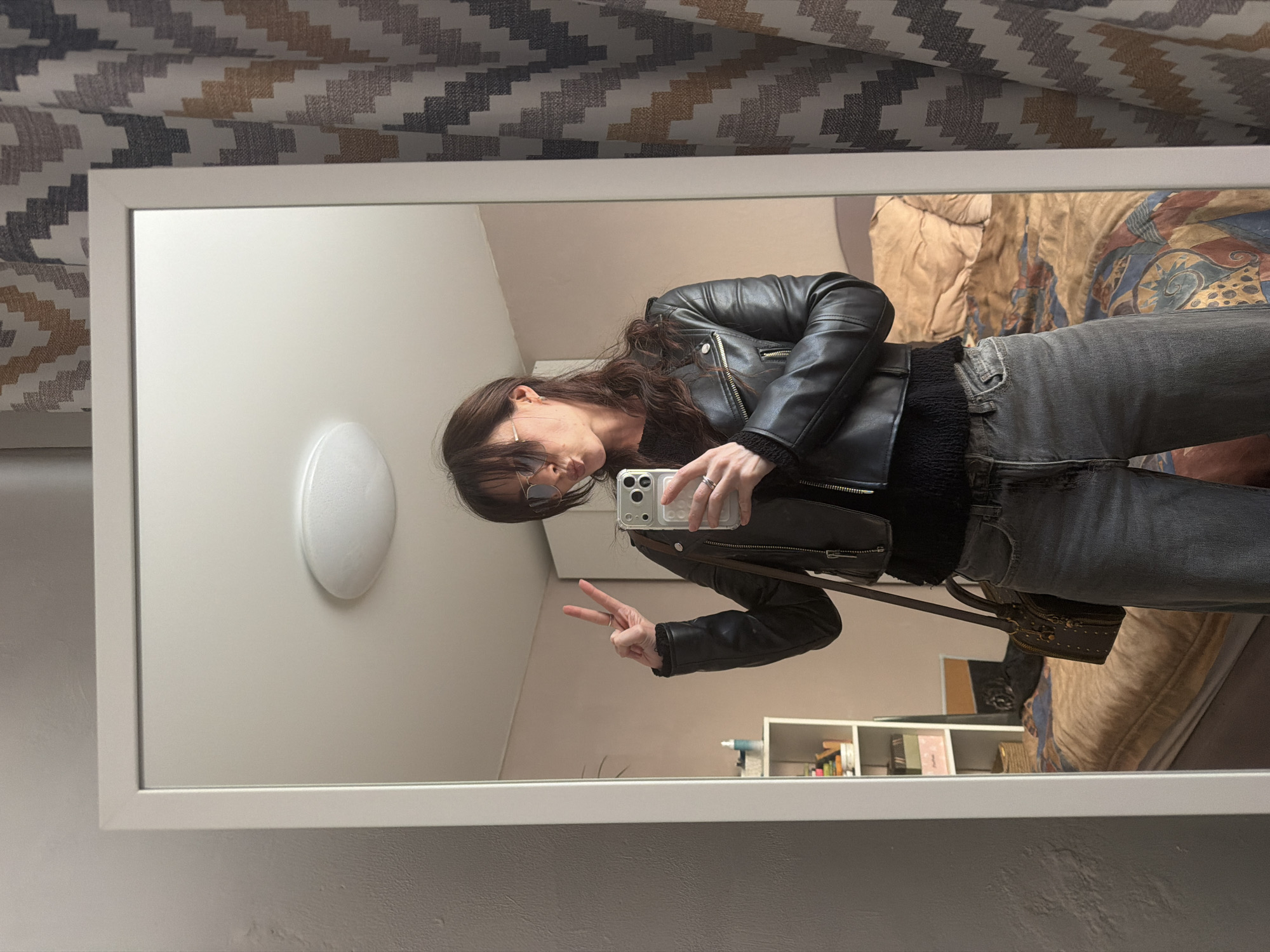 A mirror selfie shows a person wearing a black leather jacket, grey jeans, and sunglasses. The person is making a peace sign with their left hand while holding a phone in their right hand. The reflection in the mirror shows part of a room, including a light fixture on the wall, a shelf with items, and a bed with patterned bedding. The mirror is framed in white and is hung against a wall covered with patterned wallpaper. The location is implied to be in Perm, Russia.