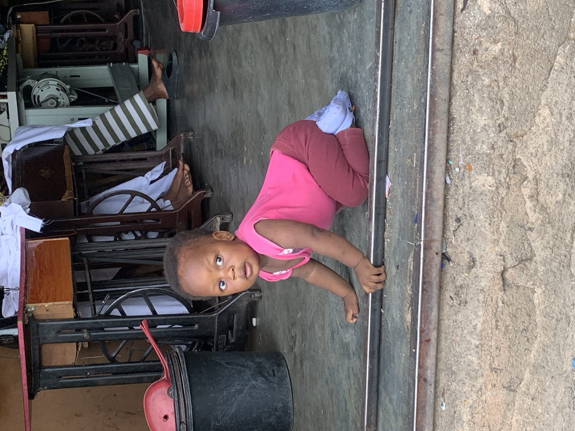 A young child, wearing a pink shirt and maroon pants with blue socks, is crawling on a concrete surface. The child is near metal rails or pipes. In the background are multiple sewing machines and tools. The location appears to be a workshop in the city of Jos, Nigeria. Another person is visible on the sewing machines.