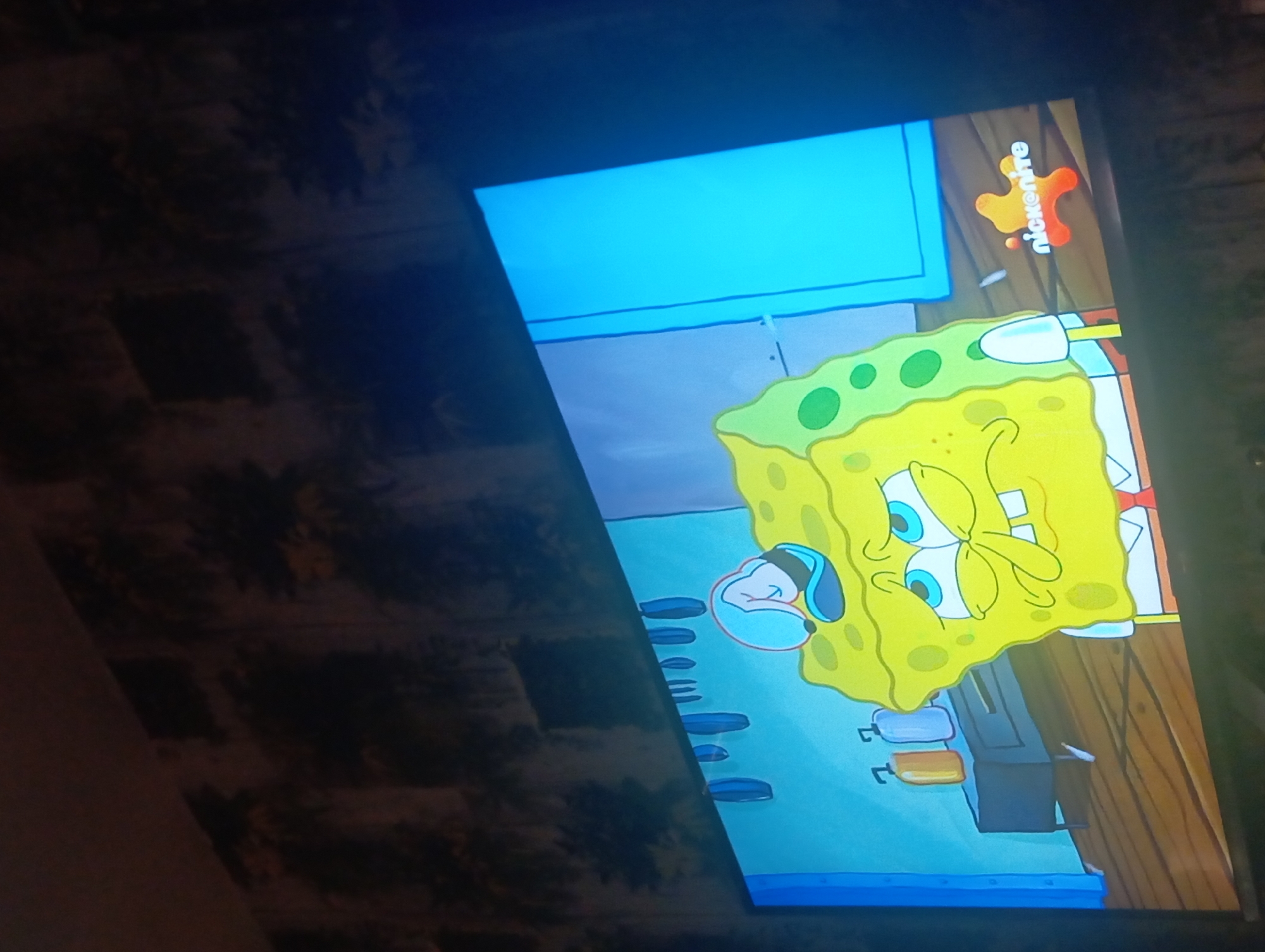 The image shows a television screen displaying an episode of the cartoon Spongebob Squarepants, featuring the character Spongebob at the Krusty Krab restaurant. The Nickelodeon logo is visible in the upper right corner of the screen. The TV appears to be located in a room with a textured wall, but the location in the city of Nkpor, Nigeria is not definitively indicated.