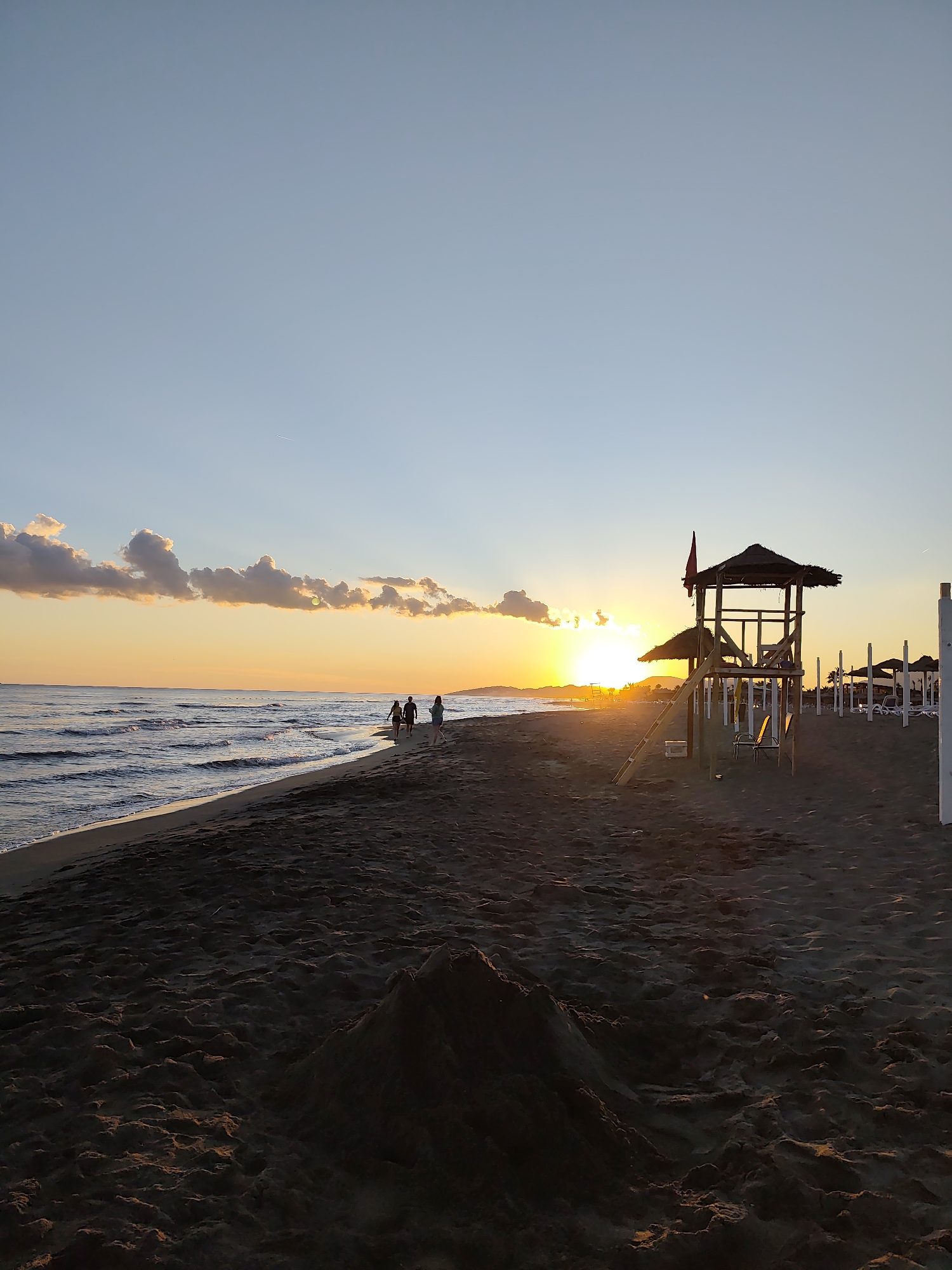 The image depicts a sunset scene on a beach in Donji Štoj - Shtoji i Poshtëm, Montenegro. The sun is low on the horizon, casting a warm orange glow across the sky and water. The sky is a clear blue with some scattered clouds illuminated by the sun's rays. The sea is calm with small waves lapping at the shore. The beach is made of dark sand and is mostly empty, with a few people walking along the water's edge. In the foreground, there is a small sandcastle. To the right, there is a wooden lifeguard tower with a thatched roof and a ladder. Further down the beach, there are several beach umbrellas and lounge chairs. The overall atmosphere is peaceful and serene.