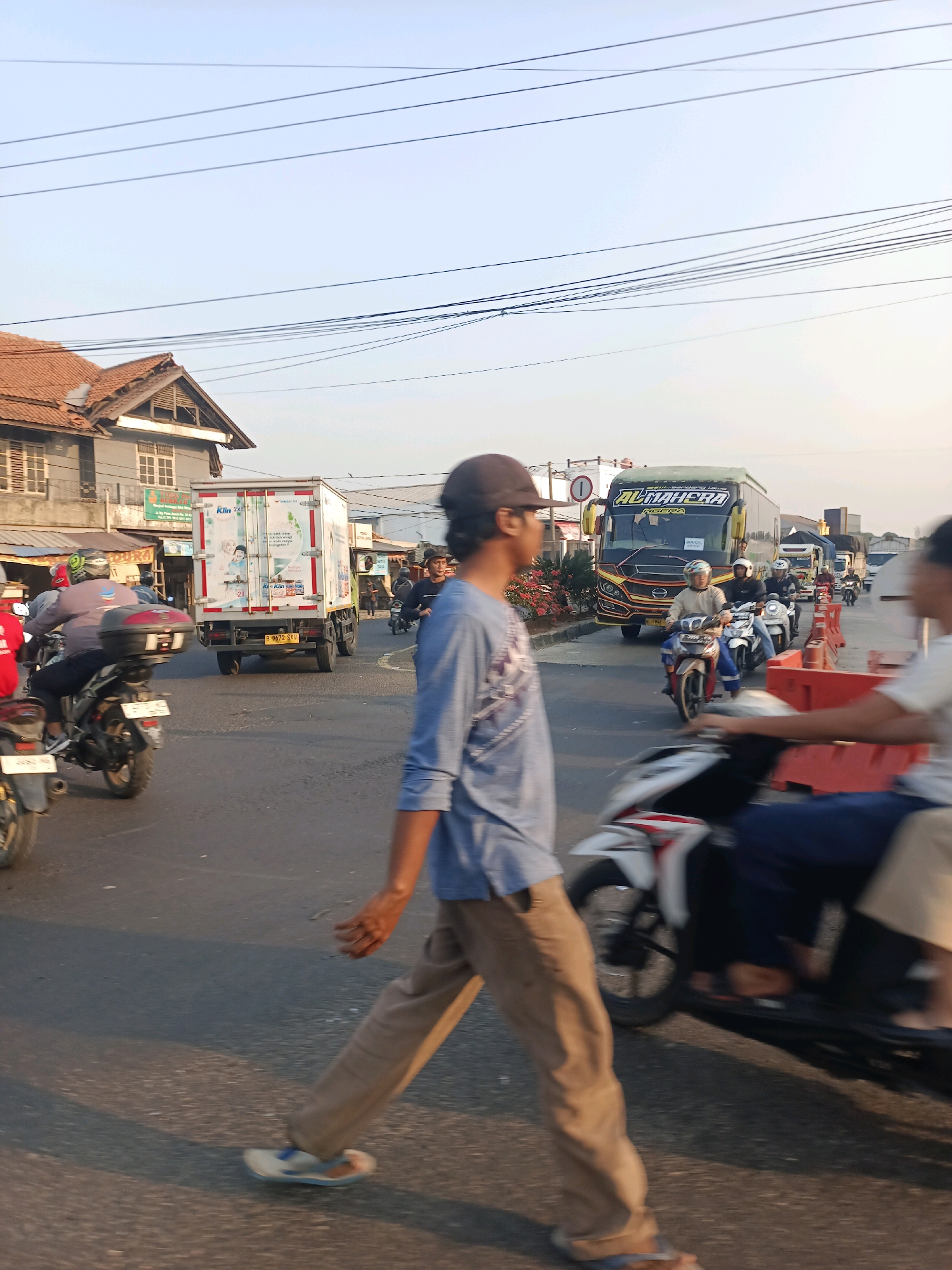 The image depicts a street scene in Karawang, Indonesia, likely during daytime given the lighting. A person, wearing a blue long-sleeved shirt and brown pants, is walking across a busy road, blurred due to motion. Several motorcycles are visible, with riders wearing helmets. In the background, a white truck with an advertisement on its side is stopped in traffic, and a yellow and black bus with the word "AL MAHERA" on it is also present. Other vehicles, including what appears to be another truck, are in the distance. The environment includes buildings on the left side of the street, possibly shops or residences, and power lines strung across the sky. Red traffic barriers are set up on the right side of the road. The overall impression is of a congested urban street with active traffic and pedestrian movement.