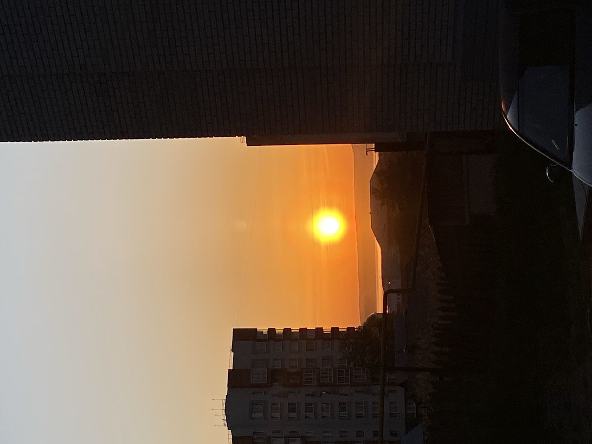 The image captures a sunset in Votkinsk, Russia. The sun is positioned centrally in the frame, radiating a bright golden light that bathes the sky in shades of orange. The view is framed by buildings on either side. On the left is a tall brick building, its texture visible in the image. On the right, a car is partially visible. In the foreground, there is an apartment block. In the background, there are hills. The overall impression is of an urban setting with a striking natural spectacle.