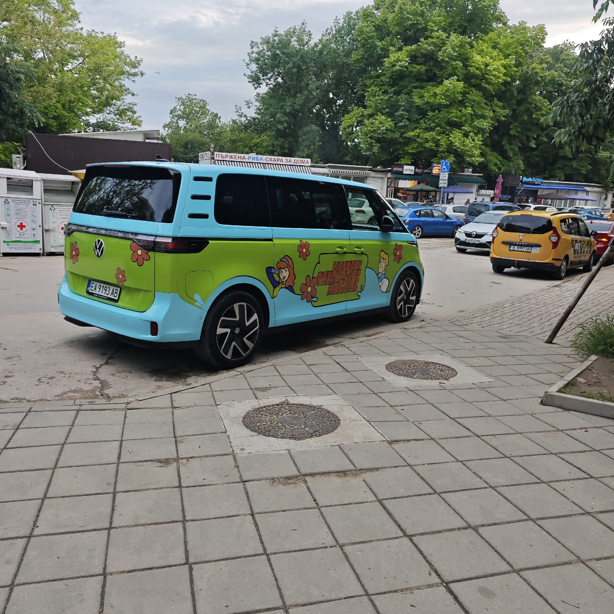 The image displays a lively street scene in Varna, Bulgaria, centered on a Volkswagen ID. Buzz van customized to resemble the "Mystery Machine" from Scooby-Doo. The van is painted in distinctive light blue and lime green, featuring red flower decals and cartoon characters, including Daphne and Fred, alongside the "THE MYSTERY MACHINE" logo on its side. Its Bulgarian license plate, "EA 9193 AB," is visible at the rear.
The photograph was taken during the day under an overcast sky. In the background, dense green foliage from large trees suggests a park or a tree-lined road. Along the street, several small commercial kiosks or shops are visible, with signs featuring Cyrillic text. Notable signs include "ПЪРЖЕНА-РИБА-СКАРА ЗА ДОМА" (Fried Fish-Grill for Home) and "НОВО" (New). Other vehicles, including a yellow Dacia Lodgy taxi with the license plate "B 8997 HH" and several other cars, are seen on the road, indicating normal urban traffic.
The foreground shows a pedestrian sidewalk made of grey square paving slabs, with two circular metal manhole covers embedded in the surface. On the far left, partially visible white recycling bins are present, one marked with a red cross symbol and text in Bulgarian related to used clothing and general household waste recycling. A blue sign with a white wheelchair symbol and the number '2' is also visible near the shops. The playful design of the van stands out against the backdrop of an everyday street environment.