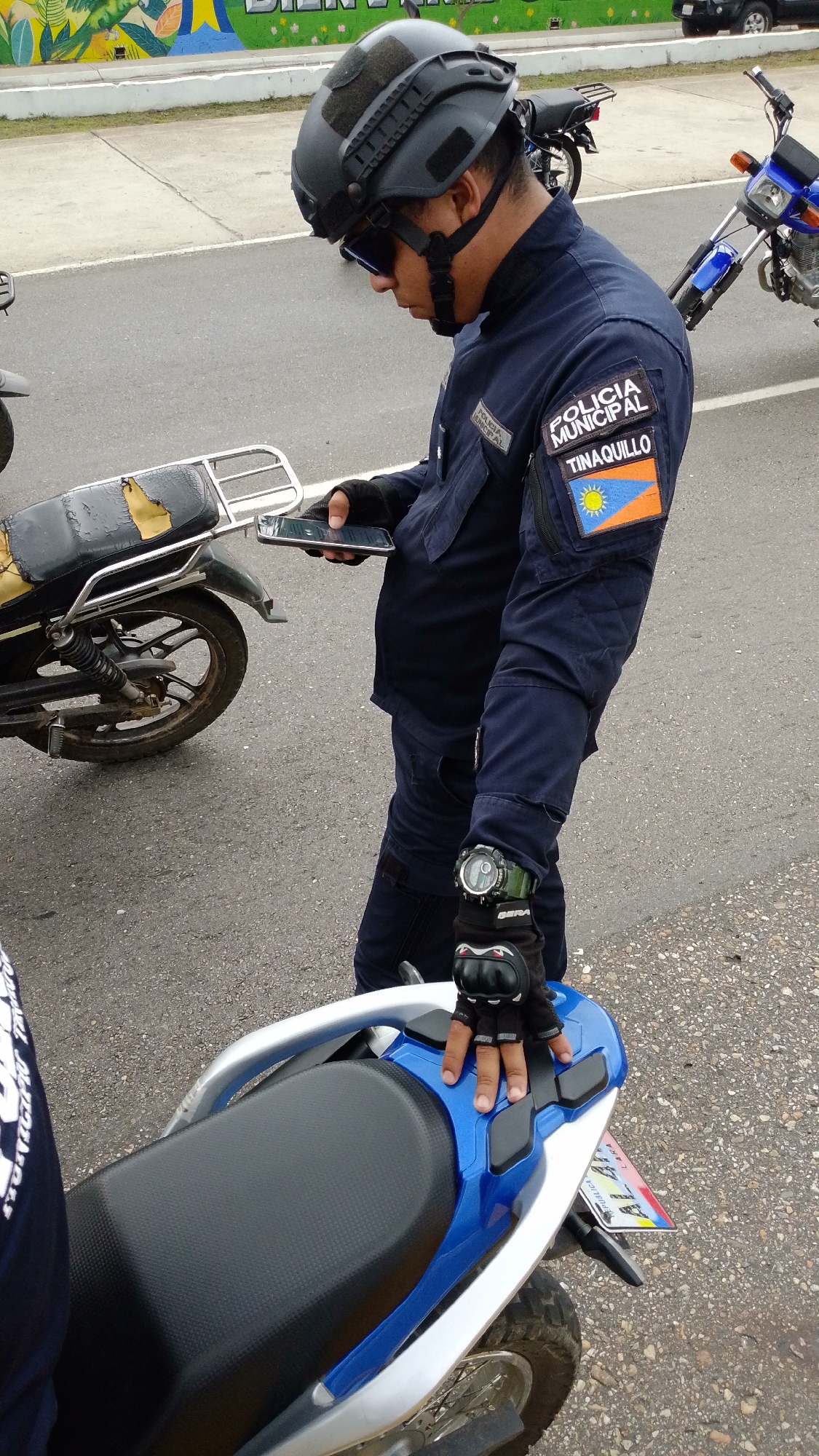 A uniformed officer of the Policía Municipal of Tinaquillo, Venezuela, stands near several motorcycles on an asphalt surface. He wears a dark navy blue uniform with a "POLICIA MUNICIPAL TINAQUILLO" patch on his left arm, a tactical helmet with goggles, gloves, and a watch on his left wrist. He is looking at a smartphone he holds in his left hand. A blue and silver motorcycle is partially visible in the foreground to his right, and two other motorcycles are visible in the background. A painted wall with colorful graphics is slightly blurred in the upper portion of the background.