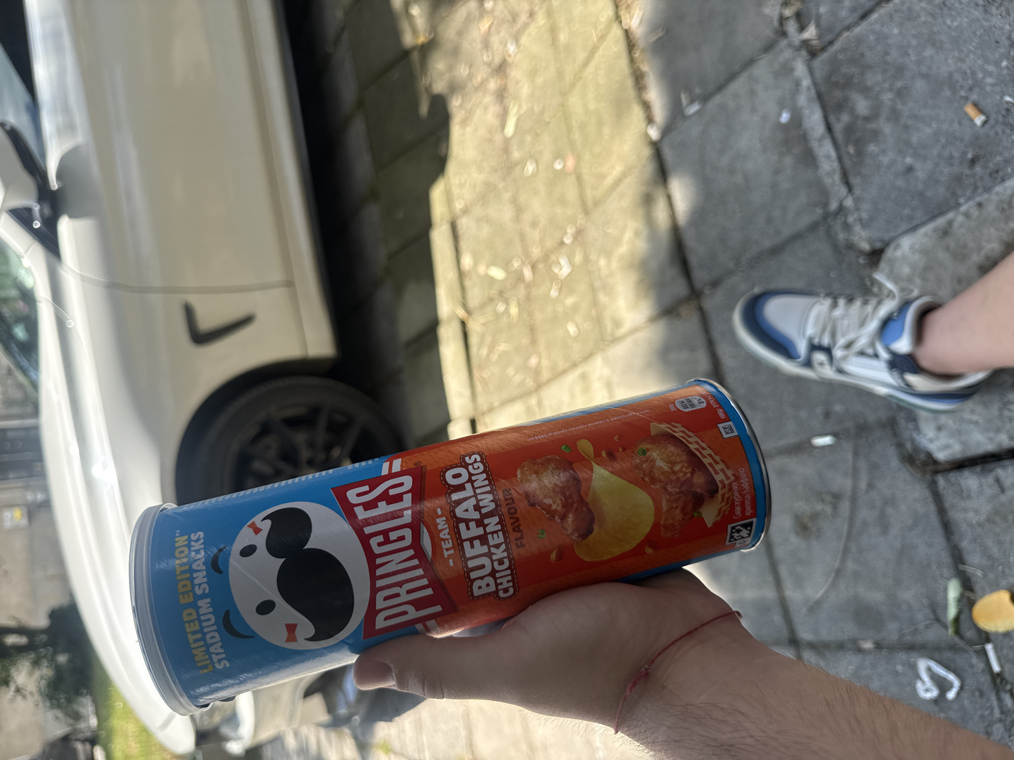 A person is holding a can of Pringles "Team Buffalo Chicken Wings Flavor" outside. In the background, a white car is parked on a paved area that appears to be a sidewalk or pedestrian zone. The ground is made of grey concrete slabs, and there is some debris on the ground, including cigarette butts and leaves. A pair of white and blue sneakers is visible in the lower right corner of the image, suggesting the person holding the Pringles is wearing them. The image has a slightly blurred quality, possibly due to movement or the camera's focus. The context of Varna, Bulgaria, is not directly discernible from the visual elements of the image itself.