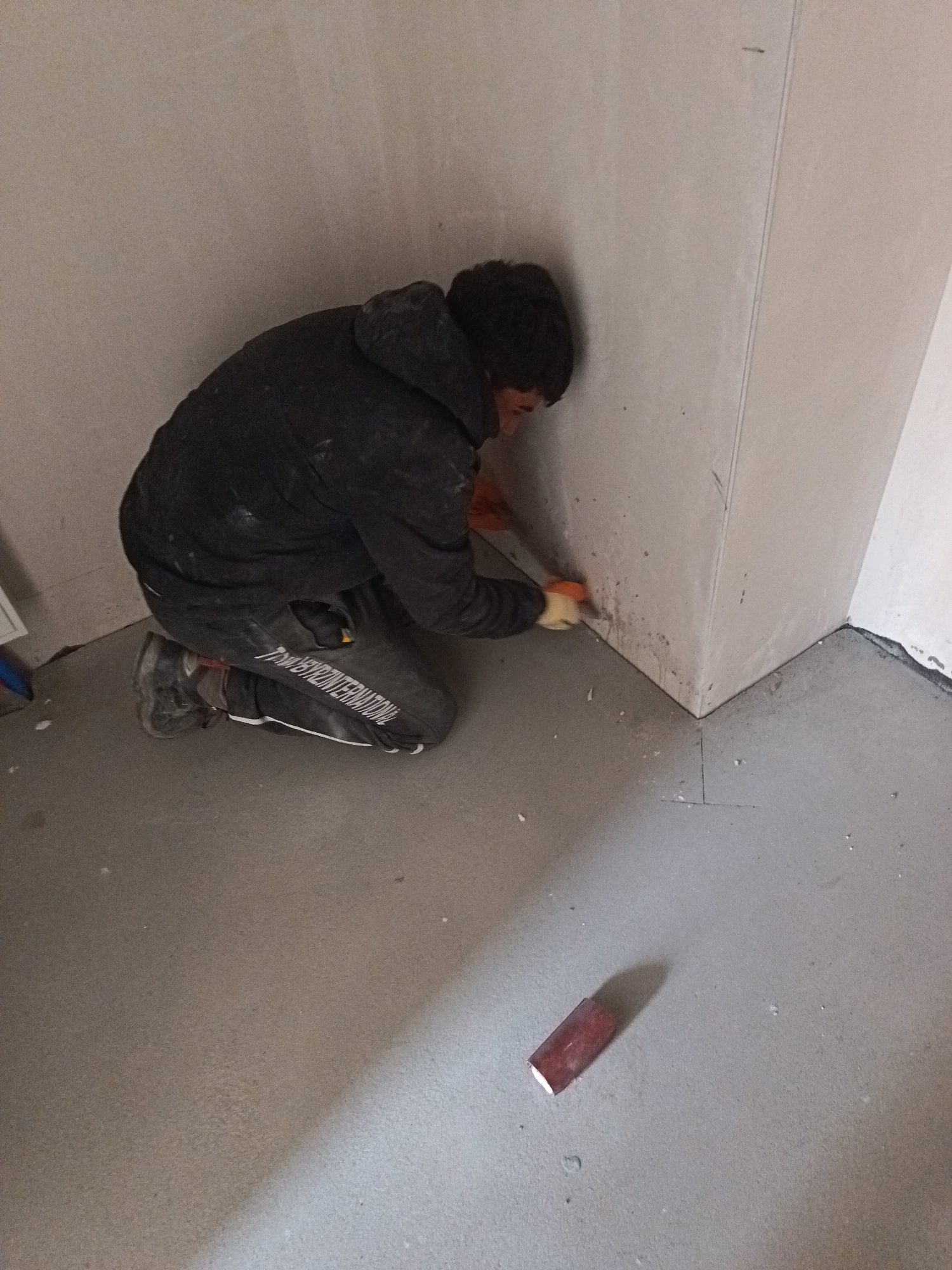 An individual is depicted kneeling on a light gray concrete or screed floor within an interior room under construction or renovation in Sofia, Bulgaria. The subject, identifiable as male, wears a dark hooded jacket, dark pants with partially visible white lettering "TRAPBYR INTERNATIONA" on the left leg, and dark work boots. Both hands are covered with orange work gloves. The individual is actively engaged in work at the junction of two unfinished light gray walls and the floor, utilizing a small tool, consistent with a scraper or trowel, held in the right hand, with the left hand positioned near the work area. Accumulations of white residue or dust are visible on the wall surface immediately surrounding the point of interaction. A distinct, reddish, rectangular object, possibly a sanding block, rests on the floor to the right of the kneeling person. The walls display visible vertical seams, suggesting panel construction or unfinished plasterwork. The floor is largely clear, with minor scattered debris.