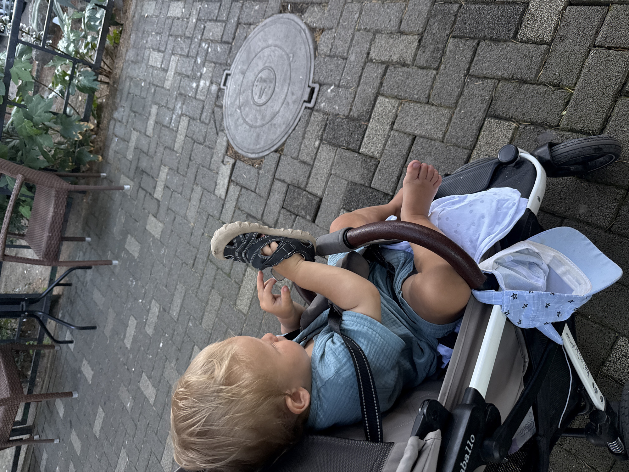 A blond-haired toddler sits in a stroller on a paved walkway. The toddler is wearing a teal romper and holding a black sandal. A blue hat with white stars hangs from the stroller. In the background, there are outdoor chairs and tables. The scene appears to be an outdoor cafe or restaurant area in Sochi, Russia.