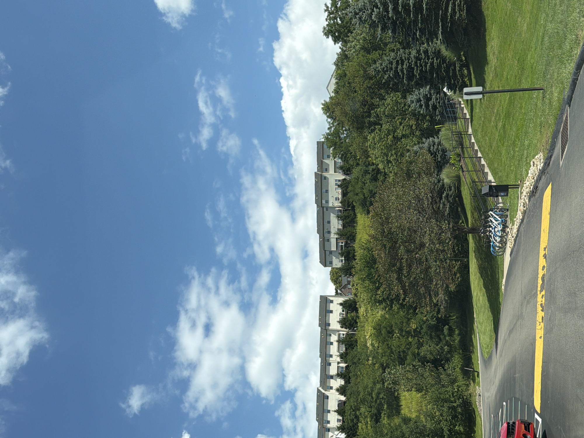The image shows a view of several townhouses on a hill, with a blue sky with white clouds above. In the foreground, there is a paved road with a yellow line. To the left, a red car is partially visible. To the right of the road, a grassy slope leads up to a set of stairs with a black railing, and a row of bicycles is parked near the bottom of the stairs. A mailbox and a sign are also present near the stairs. The townhouses are light-colored and appear to be situated in a suburban or residential area. The overall scene depicts a sunny day in a well-maintained outdoor environment.