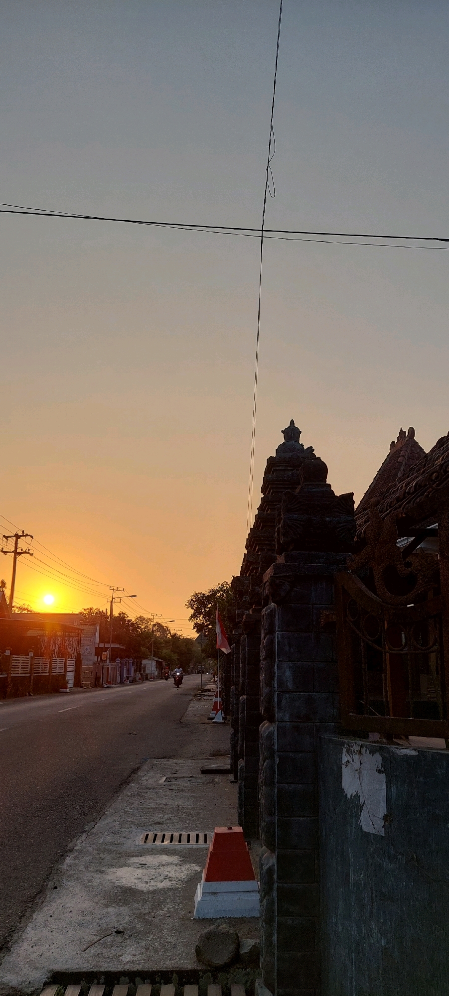 The image shows a street scene at sunset, with a vibrant orange sky dominating the upper portion of the frame. On the left, a road stretches into the distance, lined with buildings and utility poles. The sun is low on the horizon, casting a warm glow. A motorcycle rider is visible on the road. On the right side of the image, a dark, intricately carved stone fence or gate stands prominently. The fence features decorative elements and appears to be made of multiple stone blocks. A flag, possibly an Indonesian flag given the red and white colors, is mounted on a pole near the fence. The foreground shows a concrete sidewalk with a drainage grate and a small red and white marker. A thin wire runs diagonally across the sky. The overall impression is one of a quiet, possibly rural or suburban street at the end of the day.