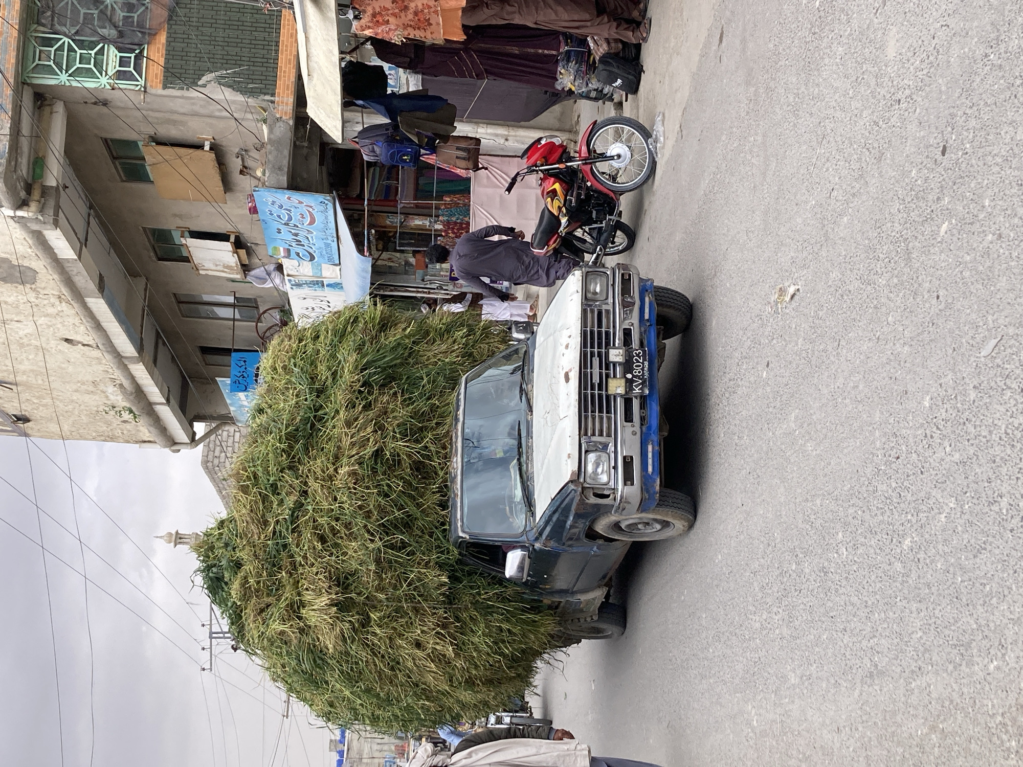 The image depicts a street scene in Quetta City Tehsil, Pakistan. A heavily overloaded blue and white pickup truck, laden with a massive mound of green grass or hay, is the central focus. A red and yellow motorcycle is parked adjacent to the truck, and a man in a dark gray outfit appears to be interacting with the motorcycle. The truck's license plate reads "KV-8023."
Clothing and textiles are hung for sale outside a building in the background. The building itself is multi-storied and features a faded blue sign with Arabic script. Above the grass mound, a minaret of a mosque is barely visible against a cloudy sky. The street appears to be paved and is lightly populated with pedestrians in the periphery.