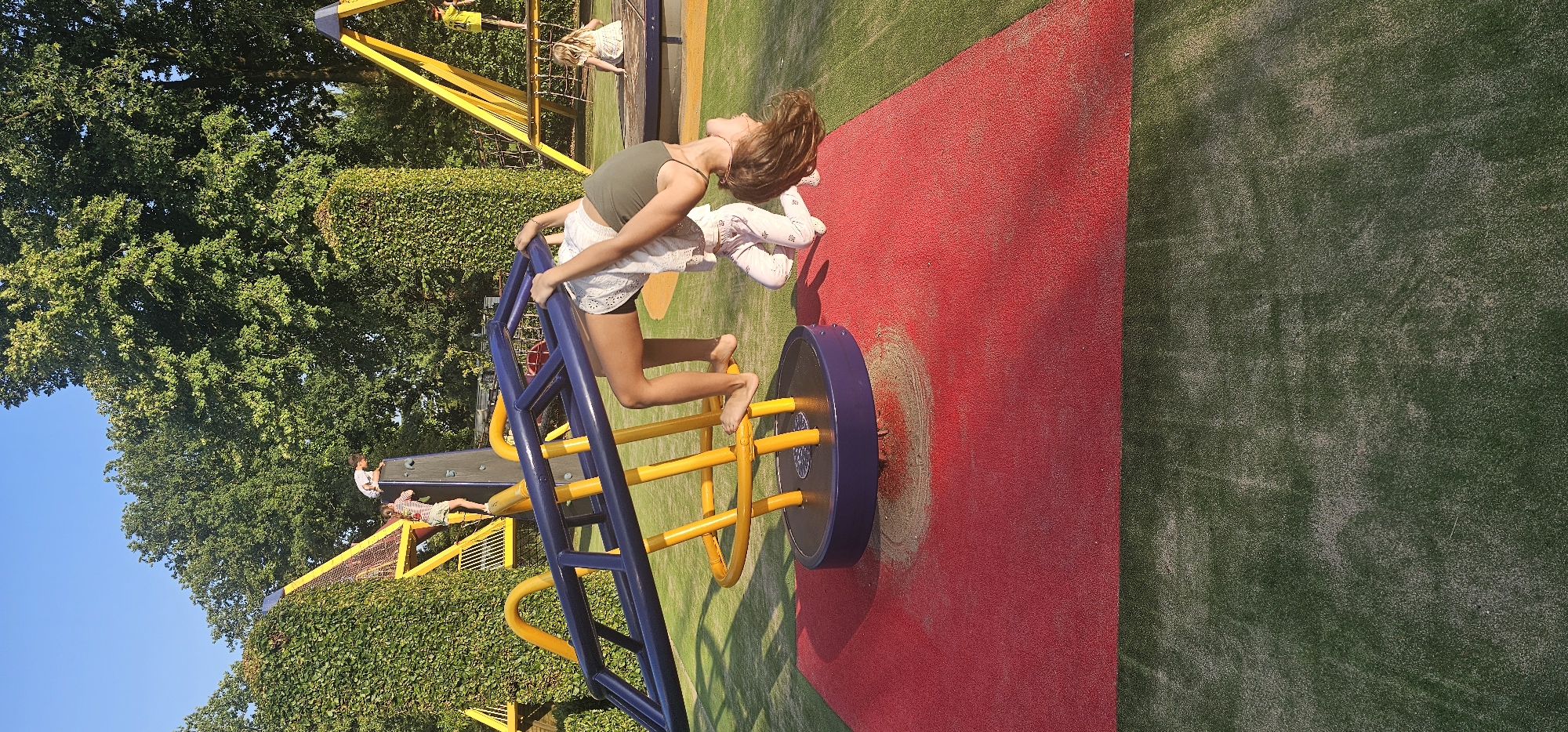 The image depicts a playground scene in Groenlo, Netherlands, on a sunny day with a clear blue sky. In the foreground, a young girl is performing an acrobatic pose on a rotating seesaw-like apparatus. She is upside down, with her arms extended, gripping the blue metal bar of the equipment. Her legs are bent, and she is wearing a green tank top and white patterned pants. Another child, possibly a younger sibling, is positioned beneath her, on their hands and knees. The seesaw is mounted on a yellow structure with a purple base, situated on a red rubberized play surface. In the background, other children are visible playing on climbing structures and a rope net. The playground is surrounded by lush green hedges and mature trees, suggesting an outdoor recreational area within the town. The ground is a combination of the red play surface and green artificial turf.