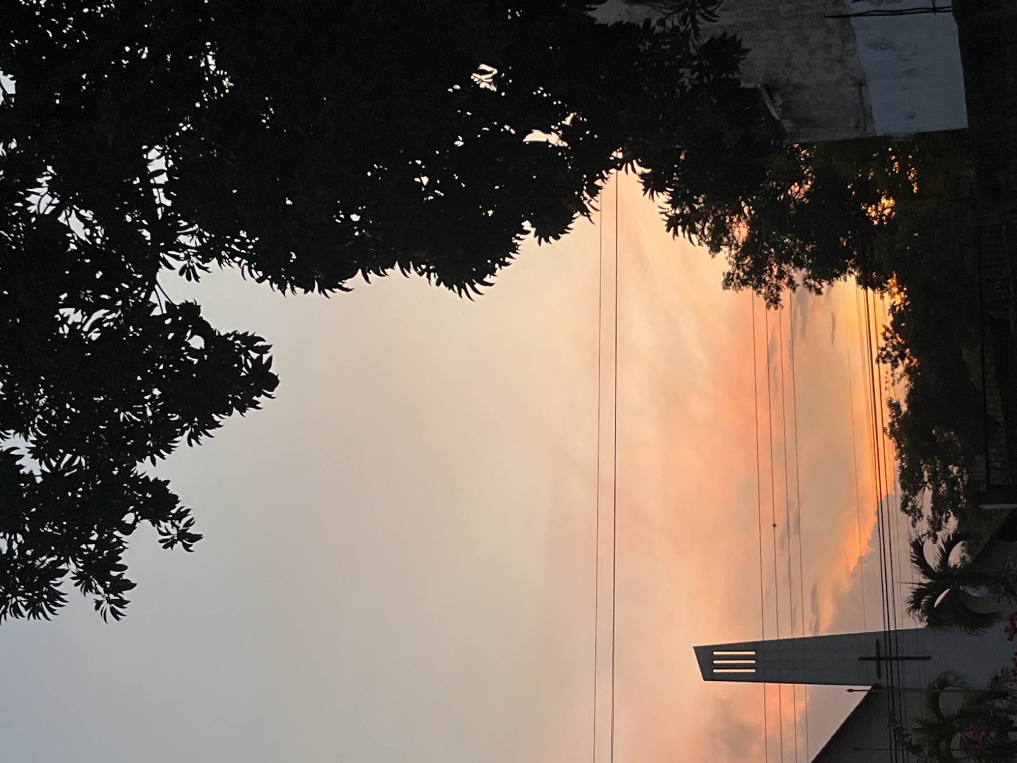 The image is taken outdoors, likely at dusk or dawn, and is oriented at an angle, suggesting the camera was held sideways. The focal point is a sky tinted in shades of orange and pink, presumably from the setting or rising sun. Silhouetted trees and foliage frame the top and sides of the view. On the right side of the frame, part of a building is visible, most likely a church, with a tall, stylized steeple or tower featuring three horizontal slits and a cross. The scene likely contains power lines. The location is Tinaquillo, Venezuela.