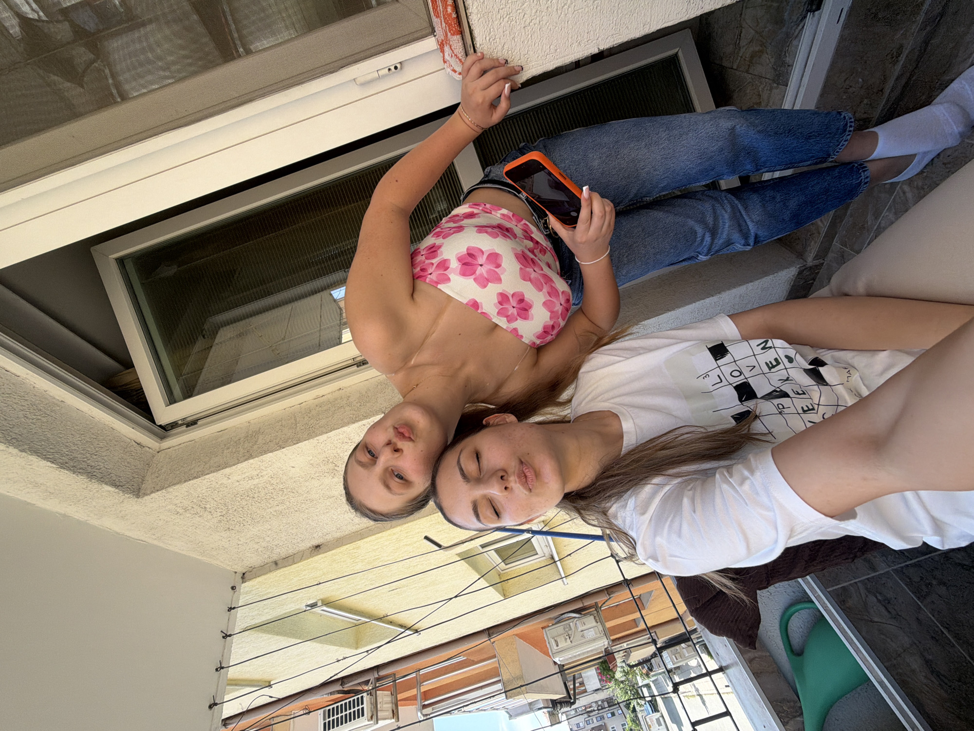 Two young women are captured in a bright, upward-angled photograph, likely taken as a selfie. The image is oriented sideways, with the women lying down and their heads pointing towards the top left.
The woman on the left (when viewed in the correct orientation after rotation) wears a pink and white floral strapless crop top and blue jeans. She holds an orange-cased smartphone in her hands, which are positioned against a textured beige wall or railing. Her eyes are open, and she has a slightly pouting expression. Her hair is light brown and long.
Next to her, the second woman wears a white t-shirt with black text and symbols arranged in a grid-like pattern. Partial text visible on her shirt includes "LOVE", "MA", "V", "P", and "RE". She has her eyes closed or nearly closed and a similar pouting expression. Her long brown hair flows around her head. She appears to be lying on a dark brown cushion or blanket on a dark tiled floor.
The background shows elements of an outdoor balcony or terrace, characteristic of urban residential areas in places like Burgas, Bulgaria. Above the women is a white window frame with a reflective glass pane and a mesh screen. Below them, visible details include a beige stucco-like wall, horizontal clothesline wires attached to wall fixtures, and a green watering can on the tiled floor. In the distance, other multi-story buildings with various architectural details are visible under a clear, sunny sky, indicating daytime.