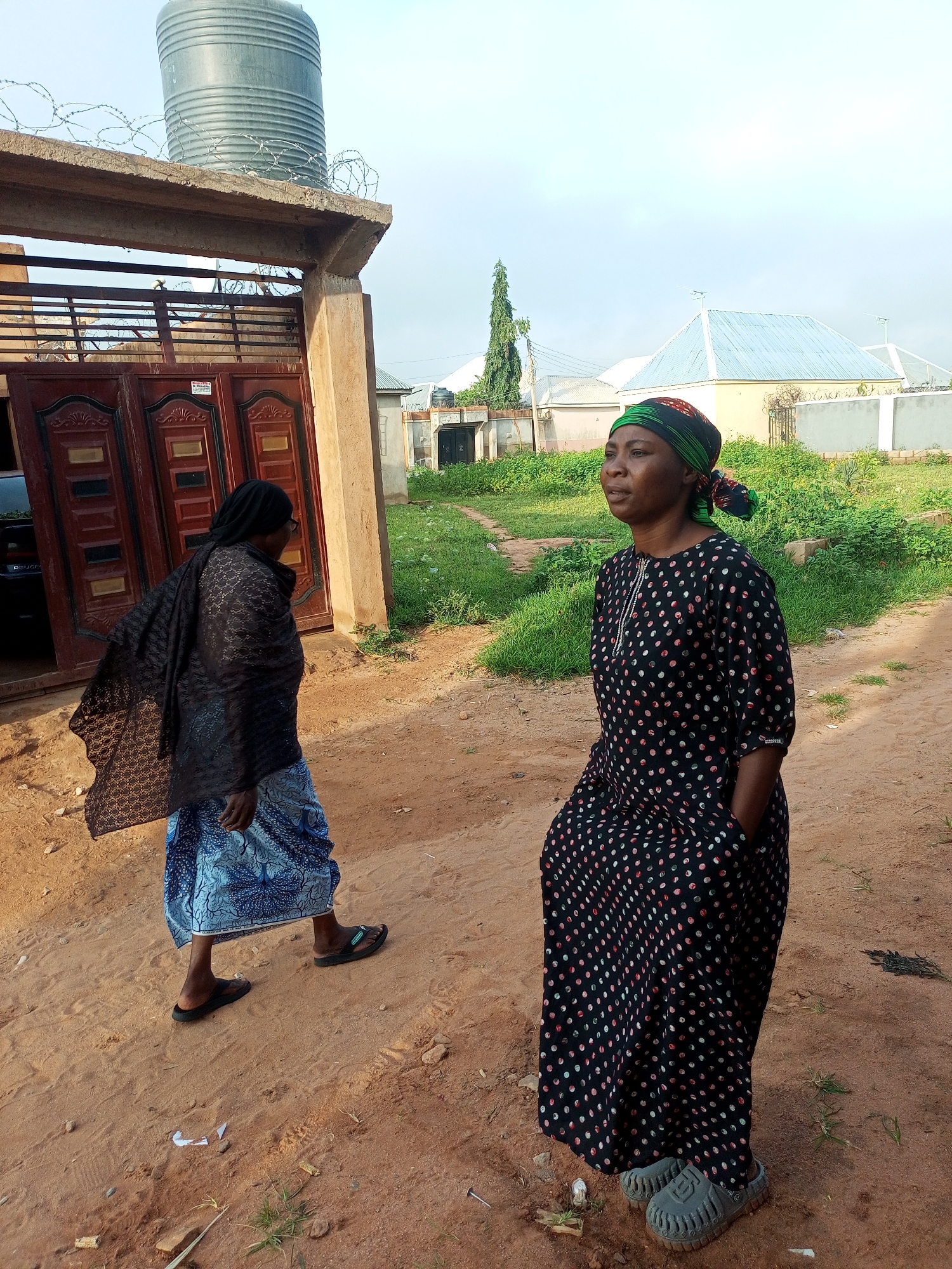 The image depicts an outdoor scene in Tempure, Nigeria. In the foreground, a woman is standing, looking to her left. She is wearing a black dress with colorful polka dots and a headscarf. Her hands are in the pockets of her dress. To her left and slightly behind her, another woman is walking away from the camera. She is wearing a dark patterned headscarf, a black patterned shawl, and a blue and white patterned skirt. She is wearing dark sandals.
Behind the women, there is a dirt path and a building with a large, ornate brown metal gate. Above the gate, there is a concrete structure with a large grey water tank on top, surrounded by barbed wire. Further in the background, there are other buildings with light-colored walls and metal roofs. Green grass and vegetation are visible throughout the scene. The sky is overcast with a pale blue hue.