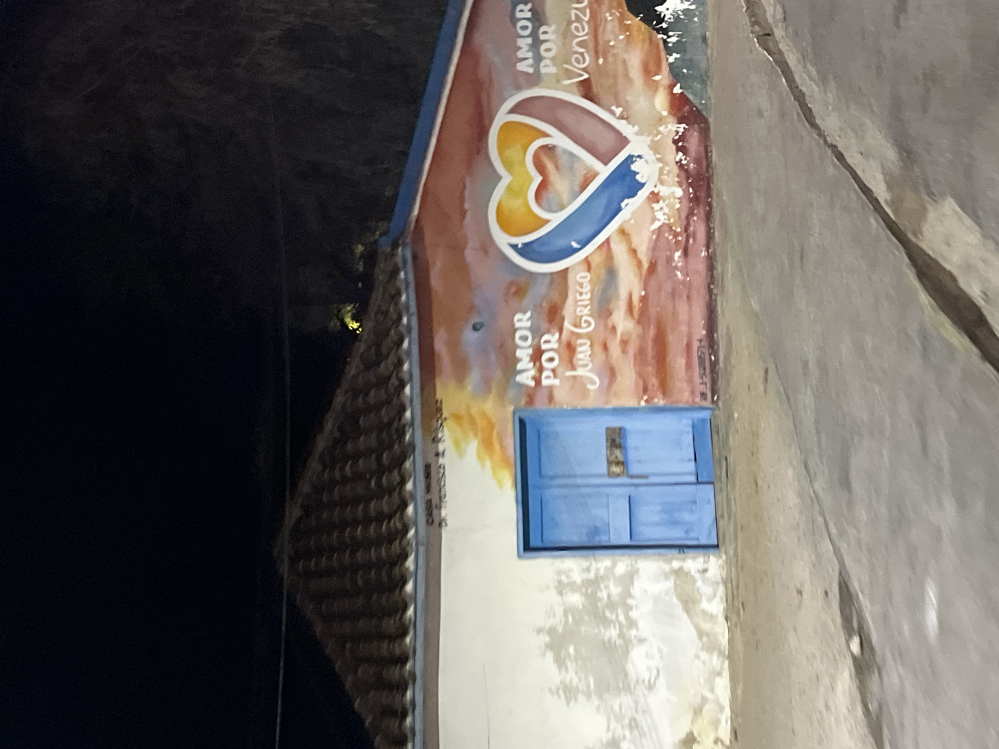 A nighttime shot shows a building in Juan Griego, Venezuela. The building has a tiled roof and a white exterior wall with a mural featuring a heart design in yellow, pink, and blue. The mural includes the phrases "AMOR POR Venezuela" and "AMOR POR Juan Griego." Below the mural is a small blue door. The wall is next to a gray sidewalk or road. The image is rotated and the scene is dimly lit. The left side of the image shows the darkness of the night sky.