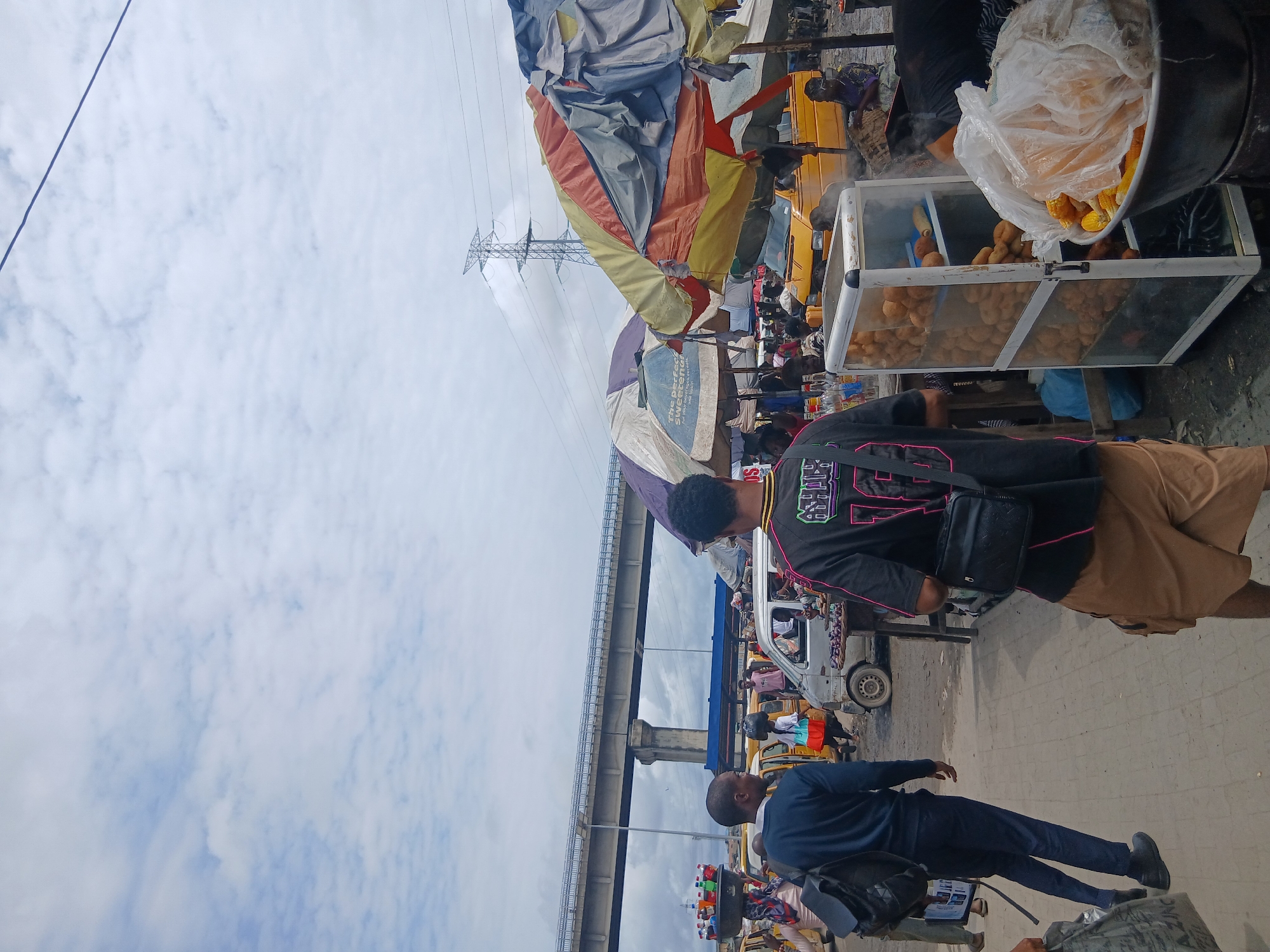 The image depicts a bustling outdoor market scene in Agani, Nigeria, under a cloudy sky. Several people are visible, engaged in various activities within the market. In the foreground, a person wearing a black t-shirt with neon pink lettering and a brown skirt walks past a food stall. The stall features a glass display case filled with fried snacks and also has corn on the cob being prepared, with steam rising from the cooking area.
Behind the food stall, a yellow minibus, typical of Nigerian public transport, is parked. More people are gathered around, suggesting a busy commercial environment. An elevated road structure with multiple lanes passes over the market, casting a shadow on the scene below. Power lines and a large transmission tower are also prominent features of the background, indicating an urban or semi-urban setting.
The overall atmosphere appears to be that of a lively market day, with many people milling about and vendors attending to their stalls. The cloudy weather suggests it could be any time of day but the lighting implies it's daytime. The presence of the minibus and the general street scene strongly point to a location in Nigeria.