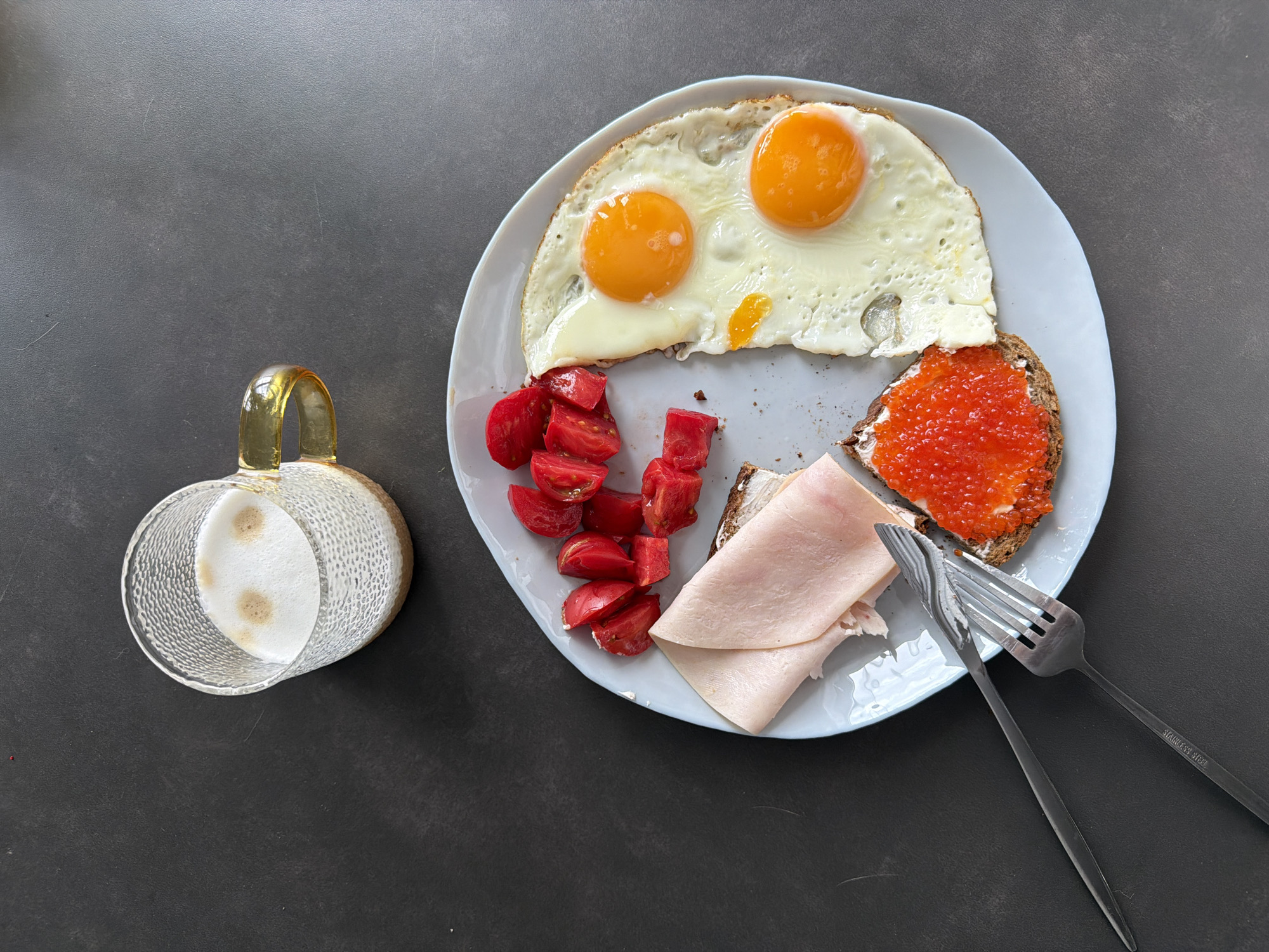 A high-angle shot depicts a plate of food alongside a beverage on a dark grey surface. The plate contains two fried eggs, diced tomatoes, slices of ham, a piece of bread with a spread of red caviar, and a knife and fork. Next to the plate is a clear glass mug filled with a foamy beverage that has a light brown spot on top. The location is Unknown, Unknown.