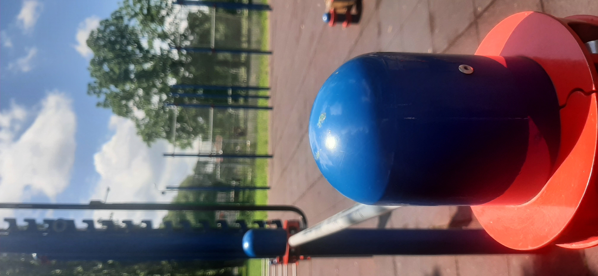 The image depicts an outdoor exercise area with a red rubberized ground surface. In the foreground, a blue, rounded cap sits atop a vertical support structure, which has a red base. A silver metal bar extends diagonally from the left side of the frame, towards the blue cap. In the background, out of focus, are multiple blue vertical poles supporting horizontal bars, typical of outdoor gym equipment. Trees and a cloudy sky are visible beyond the exercise equipment. The scene appears to be located in Yessentukskaya, Russia, as indicated by the provided information. There are no people or animals visible in the image. The overall environment suggests a public park or recreational facility.