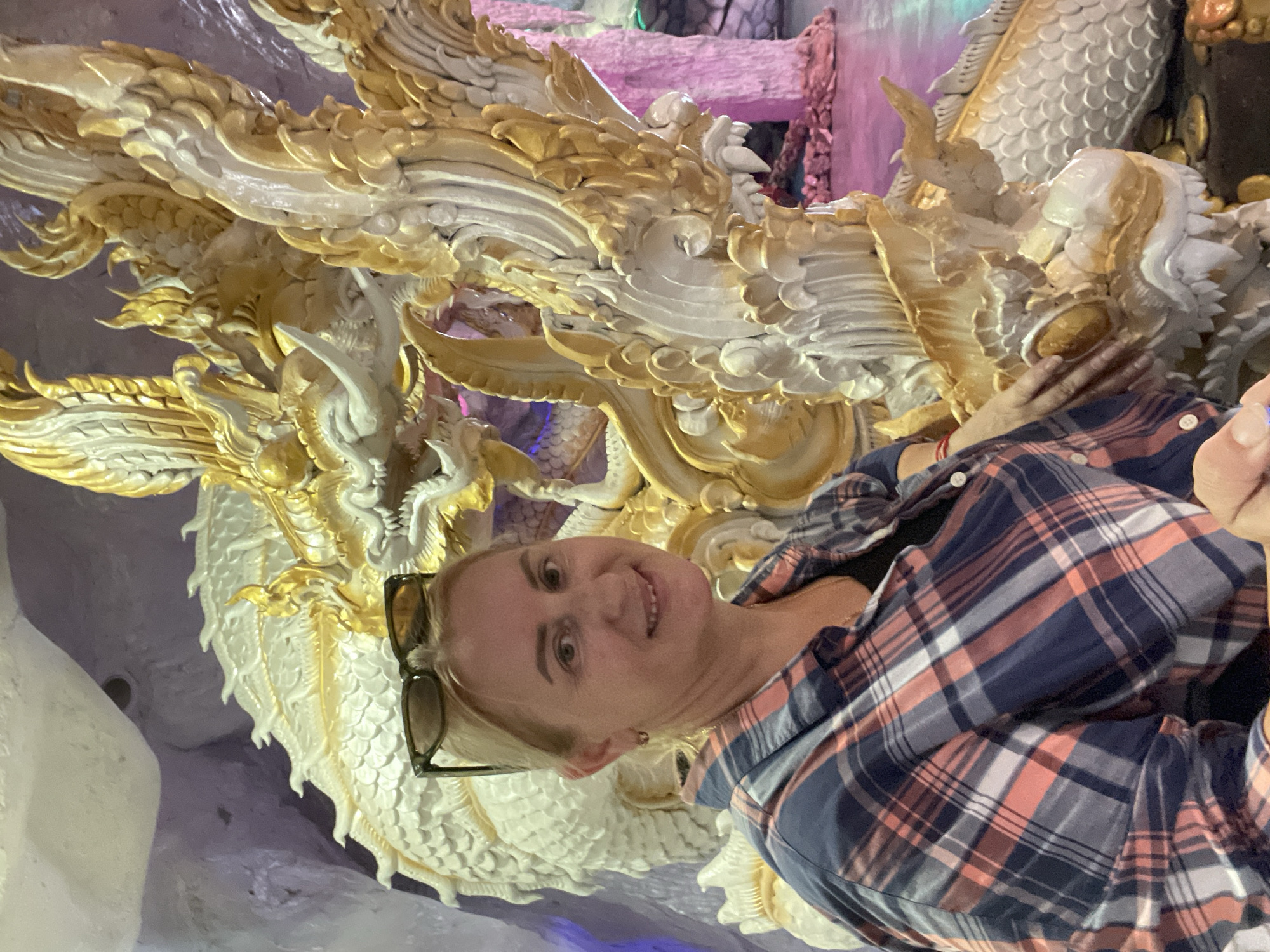 A light-skinned woman with blonde hair, wearing sunglasses on her head and a plaid shirt, smiles at the camera. She is positioned in front of a large, ornate sculpture of a dragon, which appears to be made of white and gold material. The dragon's head, with sharp teeth and golden scales, is visible to the right of the woman. The background suggests an indoor or cave-like setting with purple and pink lighting. The overall scene appears to be a tourist attraction, possibly in a temple or a themed park, given the elaborate dragon sculpture. The location is specified as Bo Win Subdistrict, Thailand.