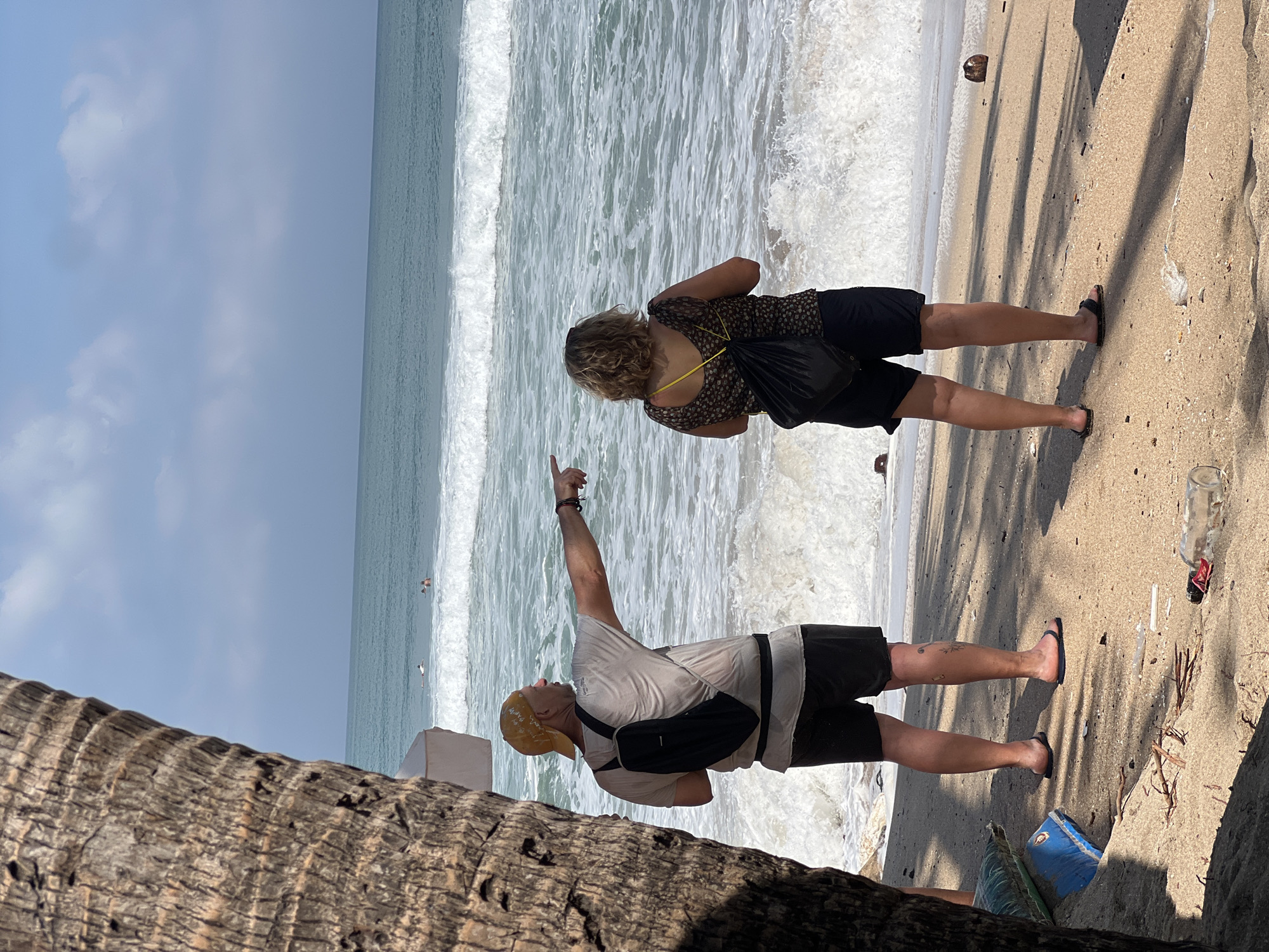 A man and a woman stand on a sandy beach in Kuta, Indonesia, facing a turquoise ocean with breaking waves under a partly cloudy sky. The man, wearing a yellow cap, grey t-shirt, and black shorts, has a black backpack and is pointing to the right. The woman, with blonde curly hair, wears a patterned dark top, dark shorts, and carries a black backpack. In the distance, two people are visible in the water, potentially surfing. The beach is littered with debris, including a plastic bottle and a coconut. A palm tree trunk is visible in the foreground on the left.