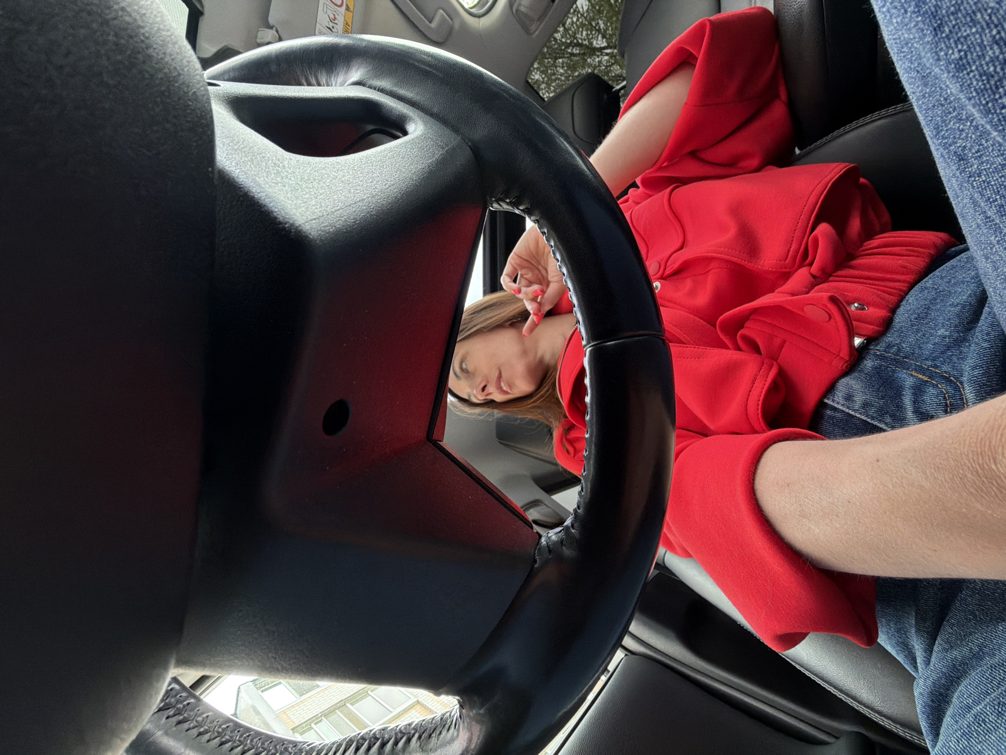 The image, taken from within a vehicle, shows a woman in a red jacket and blue jeans seated in the passenger seat. Part of the steering wheel is in the foreground, obscuring some of the scene. The woman's face is partially visible, looking towards the camera. The setting appears to be outdoors, with a glimpse of a building visible through the windshield. No specific details are discernable to indicate the location is Birobidzhan, Russia.