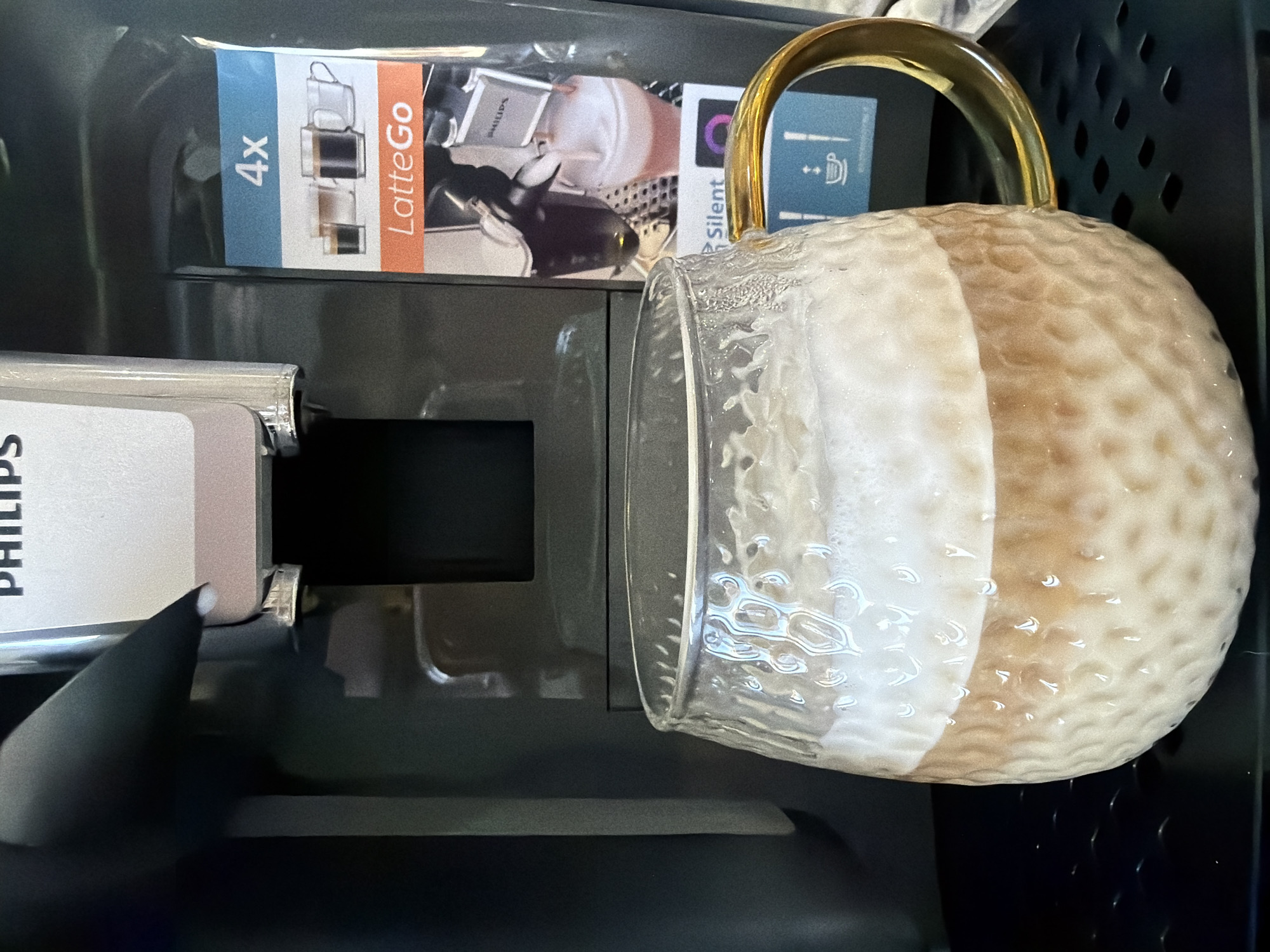 This image, captured in Chaikovsky, Russia, presents a close-up, eye-level view of a layered beverage in a clear, textured glass mug, positioned directly beneath the dispensing spout of a Philips automatic coffee machine. The mug, featuring a golden handle, contains a light brown liquid base topped with a substantial layer of white foam, consistent with a latte or cappuccino. It rests on a black, perforated drip tray. The black and silver Philips coffee machine displays a partial "PHILIPS" logo at the top. An informational sticker on the right side of the machine features text such as "4x", "LatteGo", and "Silent", along with photographic illustrations of various coffee cups and the machine's milk frothing process. No individuals are visible in the scene. The lighting indicates an indoor setting.