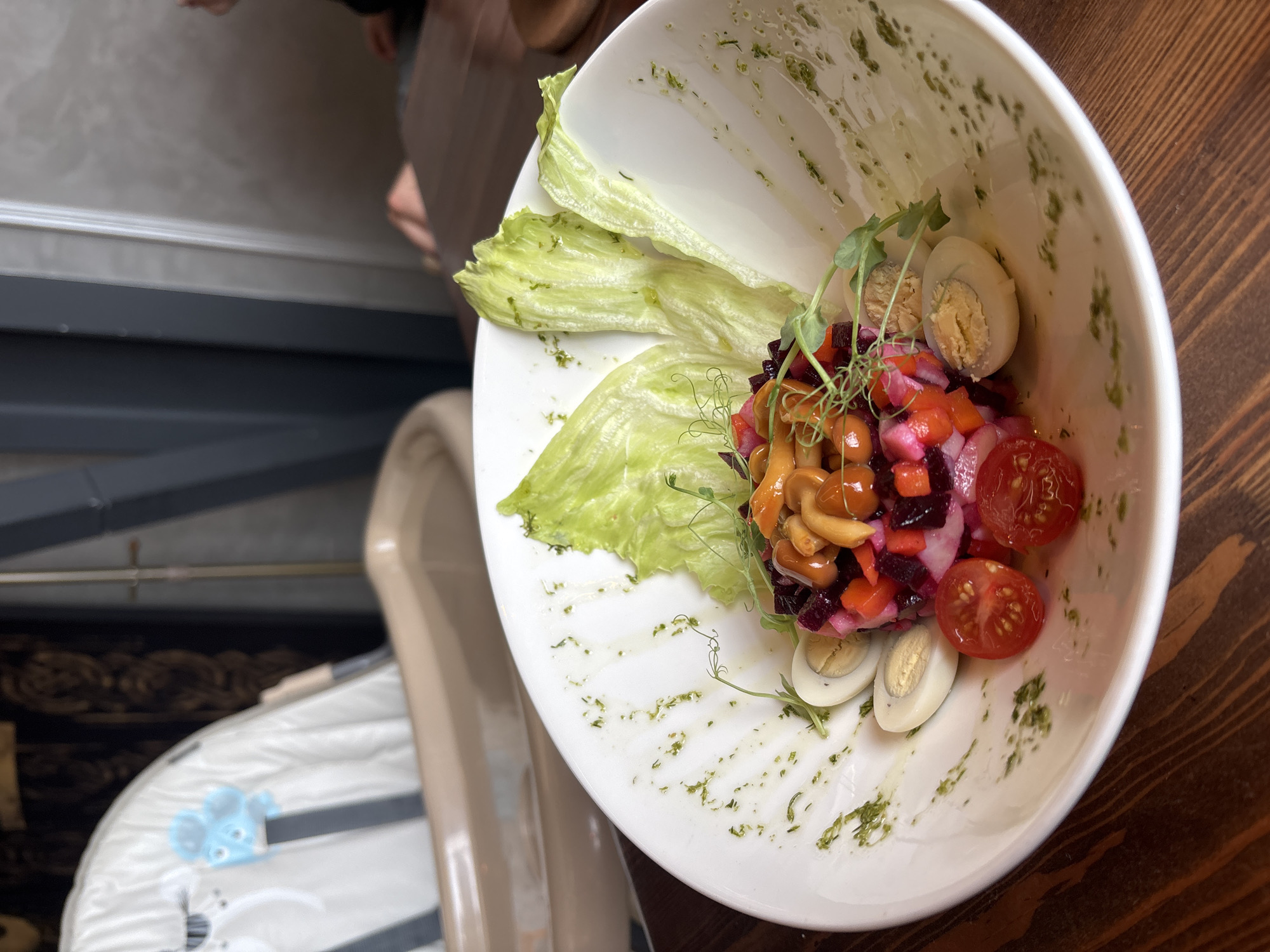 A close-up, eye-level shot shows a white bowl of salad placed on a dark wooden table. The salad is a colorful mix of diced beets, tomatoes, and mushrooms, topped with small green sprouts. Halved hard-boiled eggs are arranged around the salad. Several pieces of lettuce form a decorative border within the bowl. The background is blurred, showing a high chair, a dark wall with vertical lines, and hints of furniture. The scene appears to be in a restaurant setting in Naberezhnye Chelny, Russia.