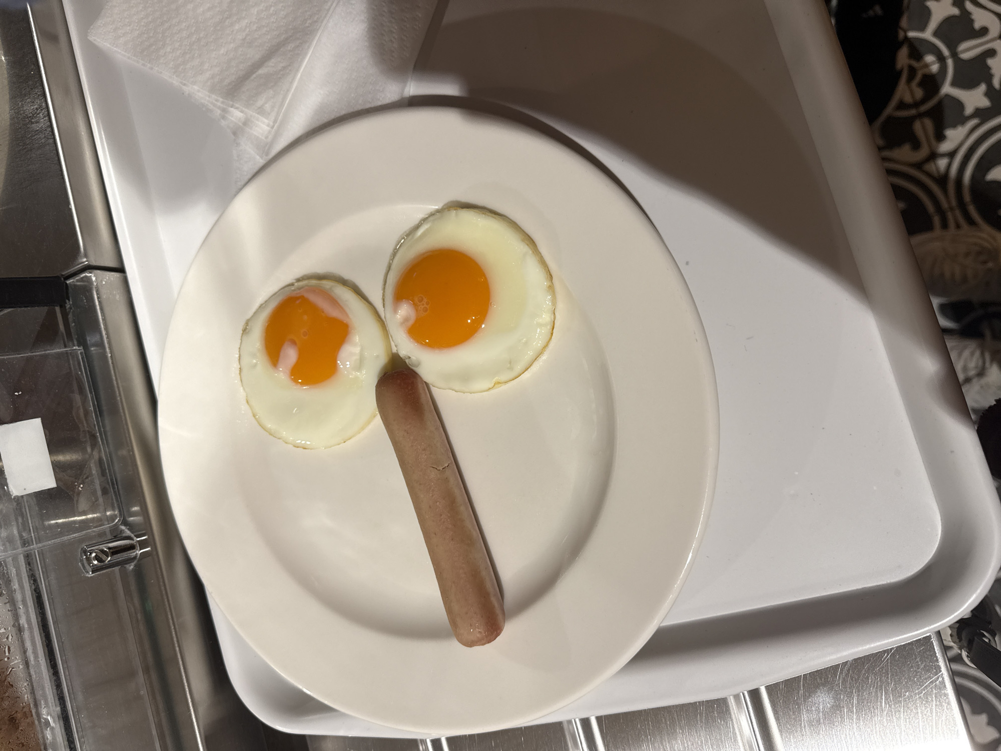 The image depicts a white plate with two fried eggs and a sausage, arranged on a white tray. The tray appears to be in a location that includes a serving area, possibly in a hotel or a similar establishment. There is no specific evidence to confirm the city of Budva, Montenegro. The setting suggests a casual dining environment.