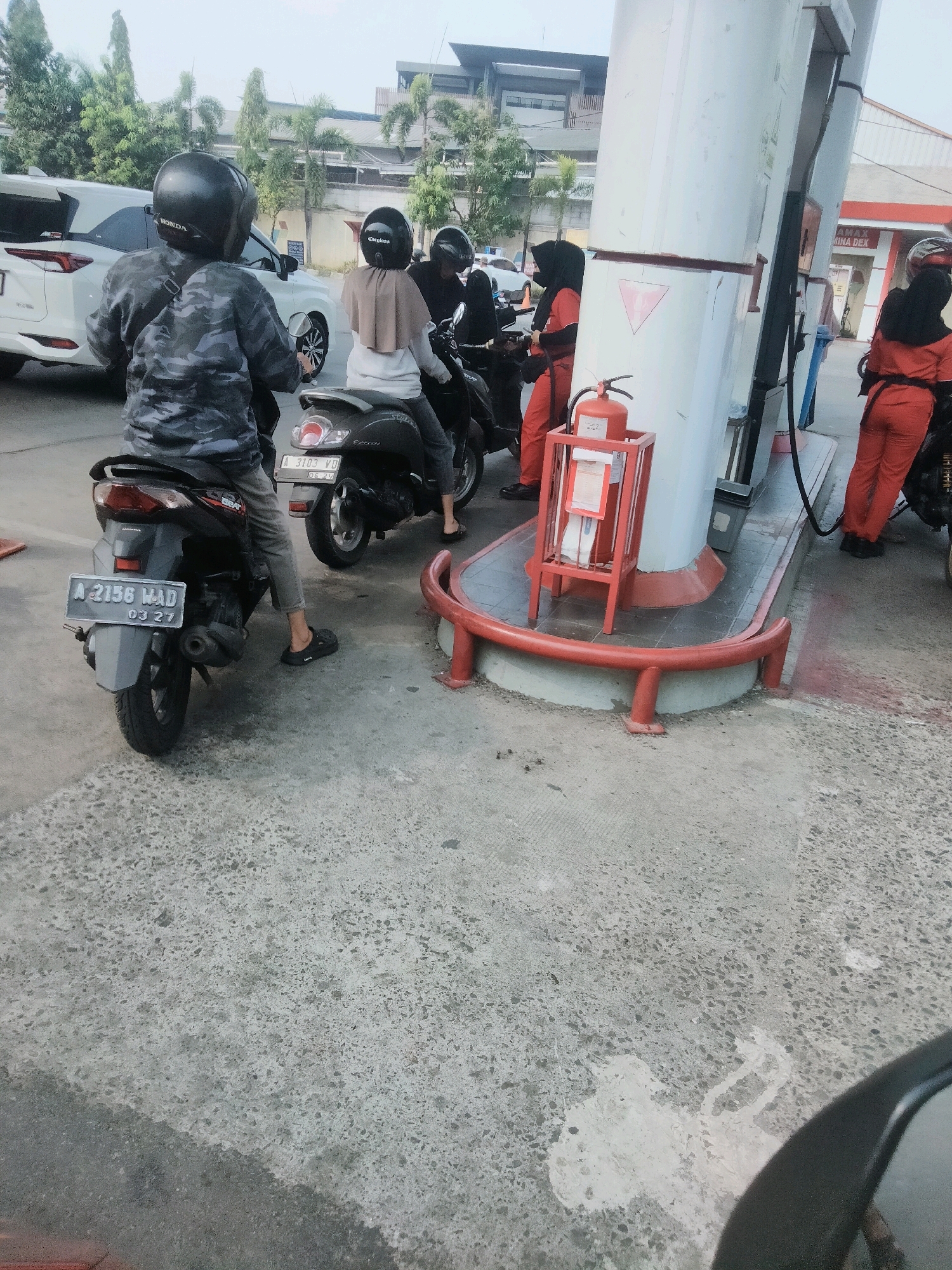 In Tangerang, Indonesia, a group of people are gathered at a Pertamina gas station. Two individuals, one wearing a camouflage jacket and helmet, and another wearing a headscarf and helmet, are seated on a black scooter with license plate number A 2156 MAD. Behind them, two gas station attendants, both in orange uniforms and headscarves, are attending to customers and refueling motorcycles. The scene takes place on a paved surface with some debris and oil stains. A white car is parked nearby, and palm trees and buildings are visible in the background.