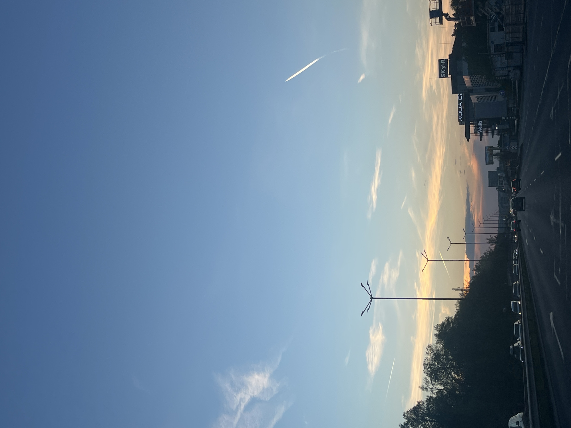 The image shows a sunset view from a road in Sofia, Bulgaria. The sky transitions from blue on the left to orange and pink hues near the horizon on the right. A contrail is visible in the upper left portion of the sky. Streetlights line the road, and dense trees are near the road. Cars are driving on the road. Billboards for businesses such as SKY-R and Aqua City are along the road.