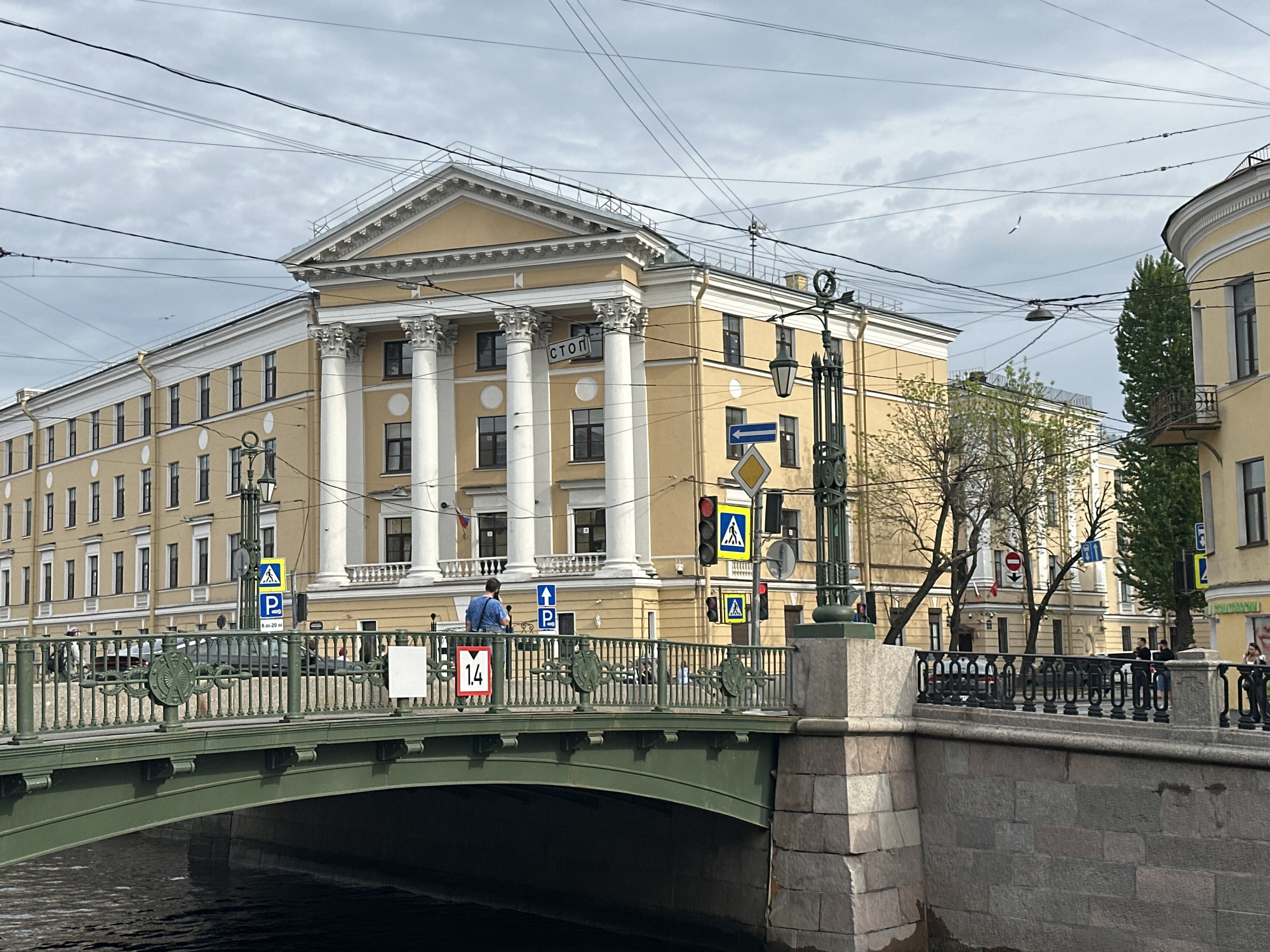 The image shows a view of the Tauride Palace from across the Fontanka River in Saint Petersburg, Russia. A green bridge spans the river in the foreground. A man in a blue shirt stands on the bridge, looking to his left, partially obscuring a red-and-white sign that reads "1.4." Street lamps and traffic signs are visible. The river surface is dark and still. The sky is overcast. Overhead power lines are visible.