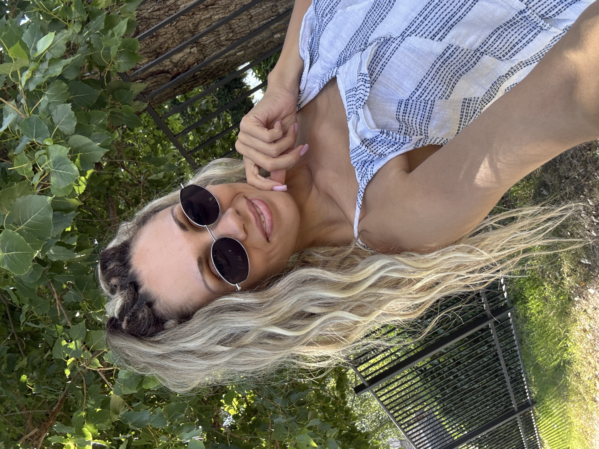 A woman with blonde, wavy hair pulled into a high ponytail is pictured outdoors. She is wearing round, dark-lensed sunglasses and a white and blue striped off-the-shoulder top. Her right hand is resting on her chin, and she is smiling. In the background, green foliage from a tree and a black metal fence are visible. The image appears to be taken in Rusanovo, Russia, as part of a geotagged set of images.